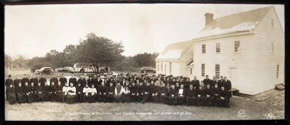 A large group of men and women seated in front of a white house, with old cars parked in the background, taken in the early 1900s.