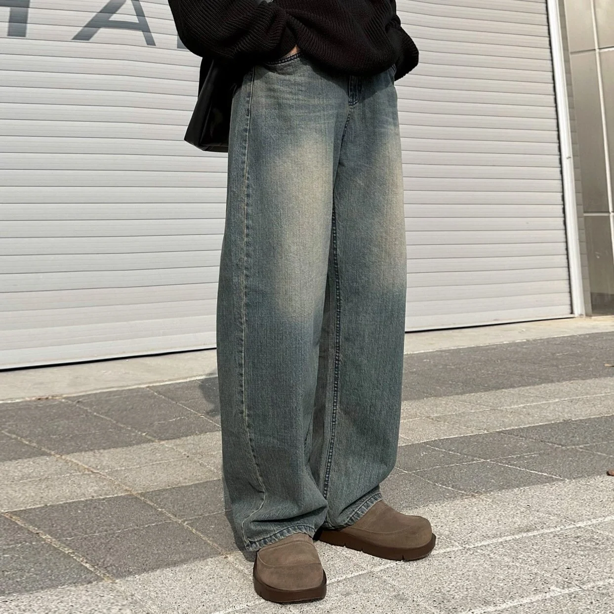 Person wearing wide-leg jeans, a black sweater, and brown boots standing outside in front of a metal garage door.