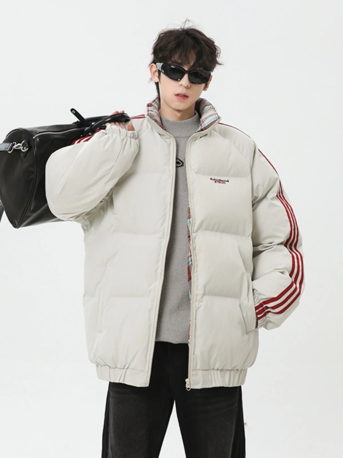 A young man wearing sunglasses, a beige puffer jacket with red stripes on the sleeves, and holding a black duffel bag over his shoulder, standing against a white background.