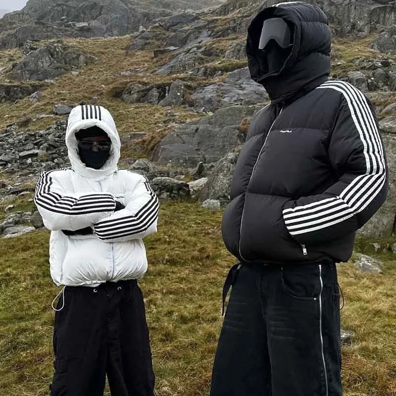 Two people wearing black and white puffer jackets with track stripe details, hooded and face masks, standing outdoors on grassy terrain with rocks and a hillside background.