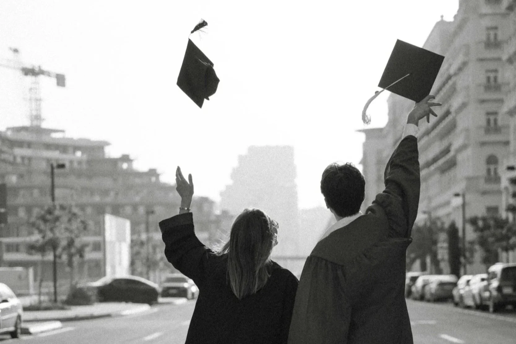 a black and white photo of a man and woman wearing graduation robes throwing up their graduation caps in the air