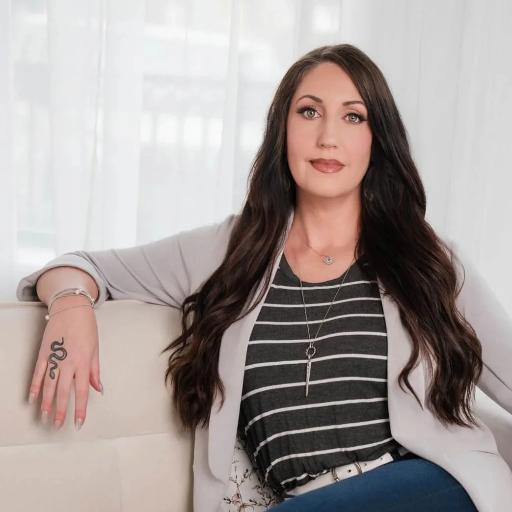 a headshot of megan stewart wearing a striped shirt, blazer, and sitting on a couch