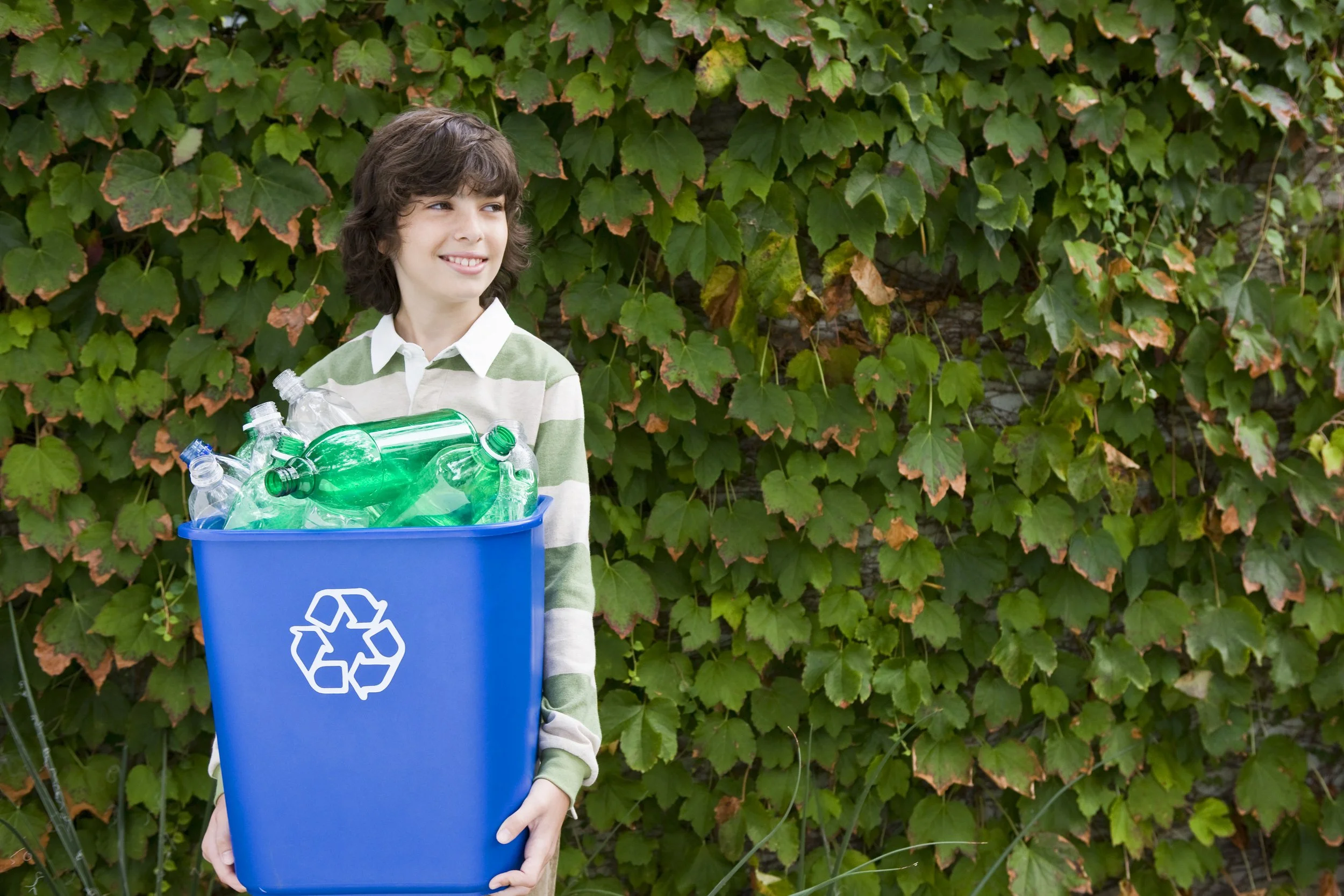 A boy with dark, curly hair holding a blue recycling bin filled with empty plastic bottles in front of a leafy green wall.