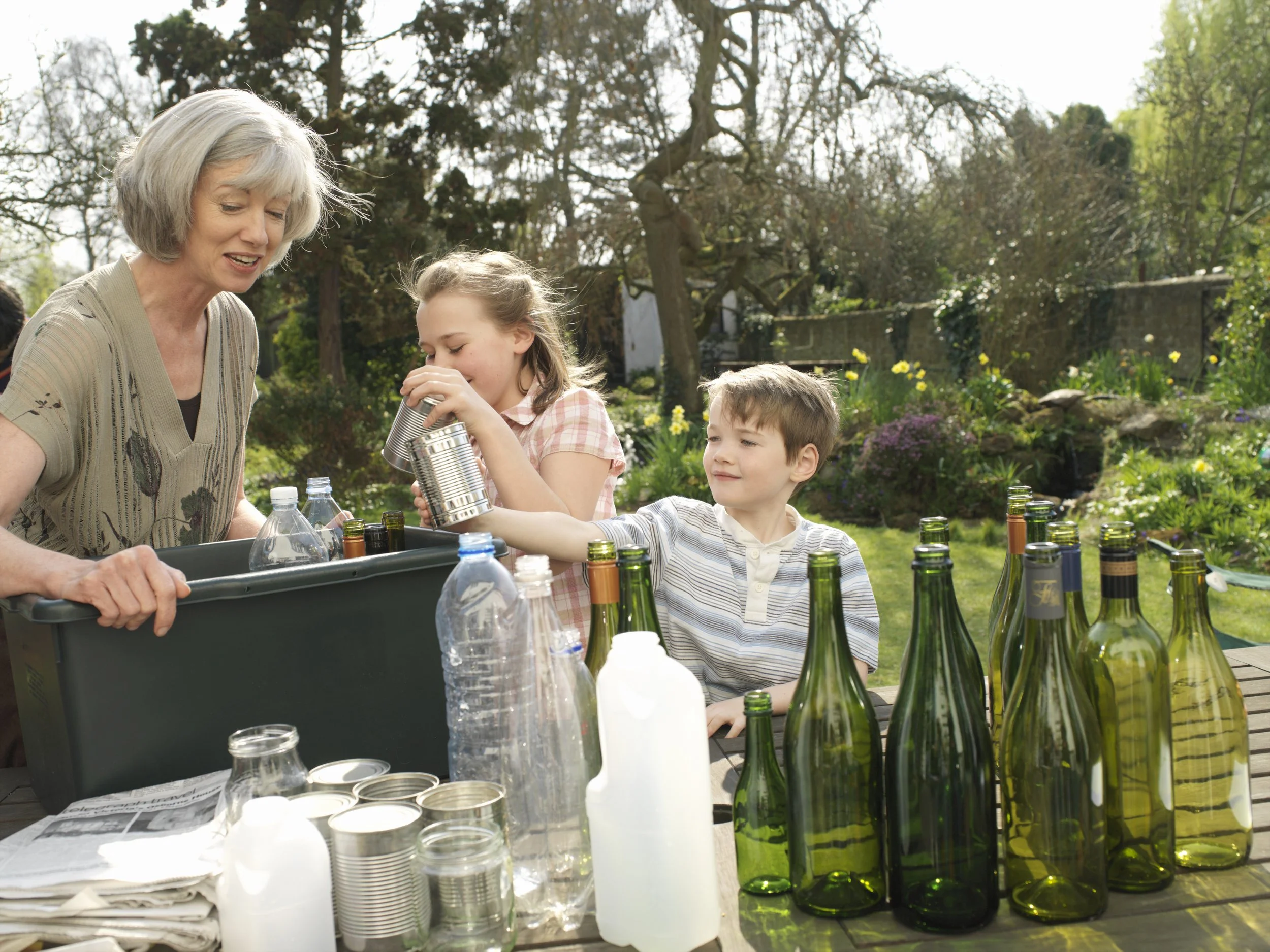 A group of children and an elderly woman outdoors surrounded by trees and flowers, engaging in an activity involving bottles, cans, and other containers on a table.