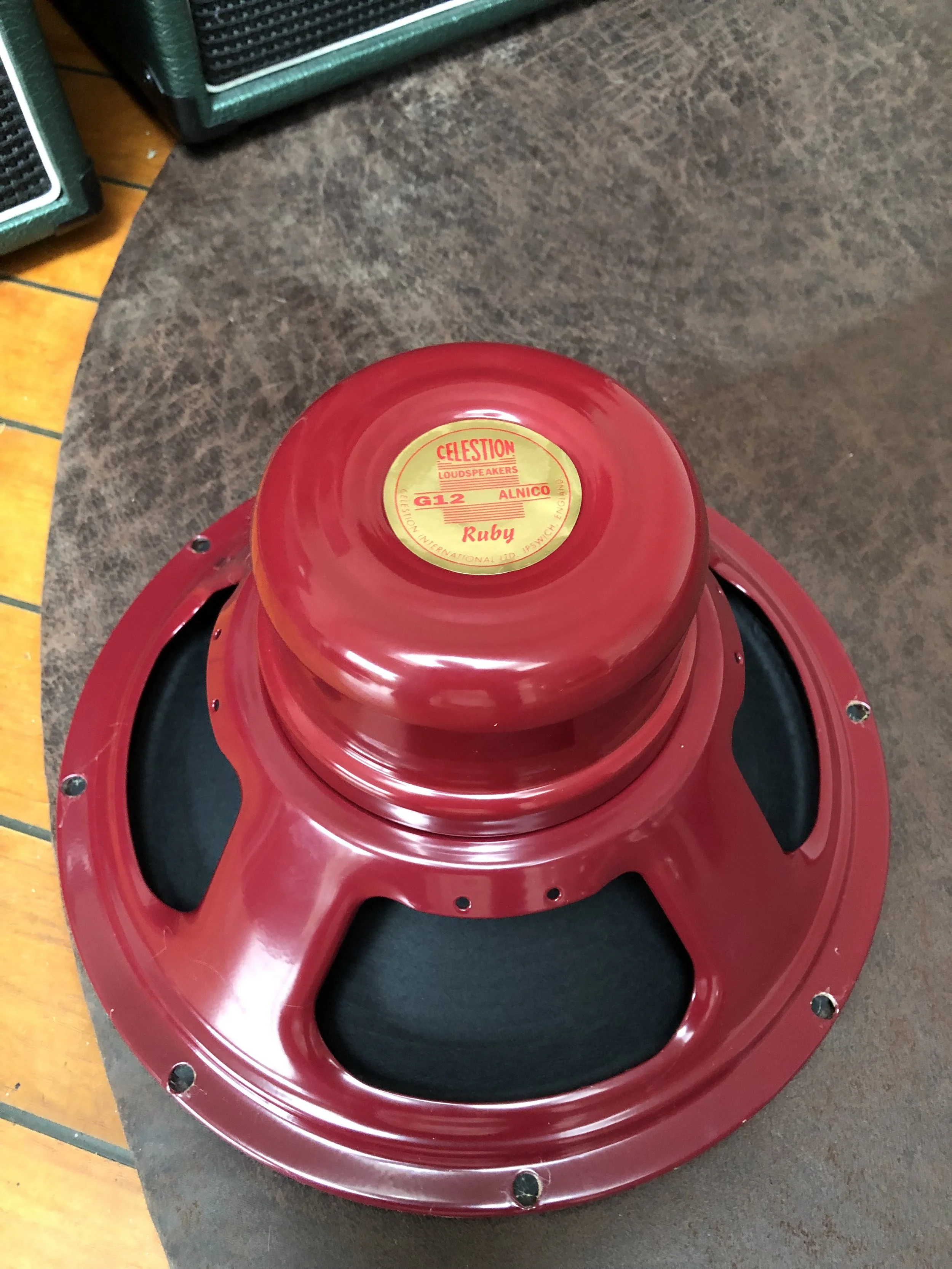 Un haut-parleur Celestion Alnico Ruby  de 1é pouces, placé à l'envers sur une surface en bois. La tache centrale est une étiquette jaune avec du texte rouge et noir.