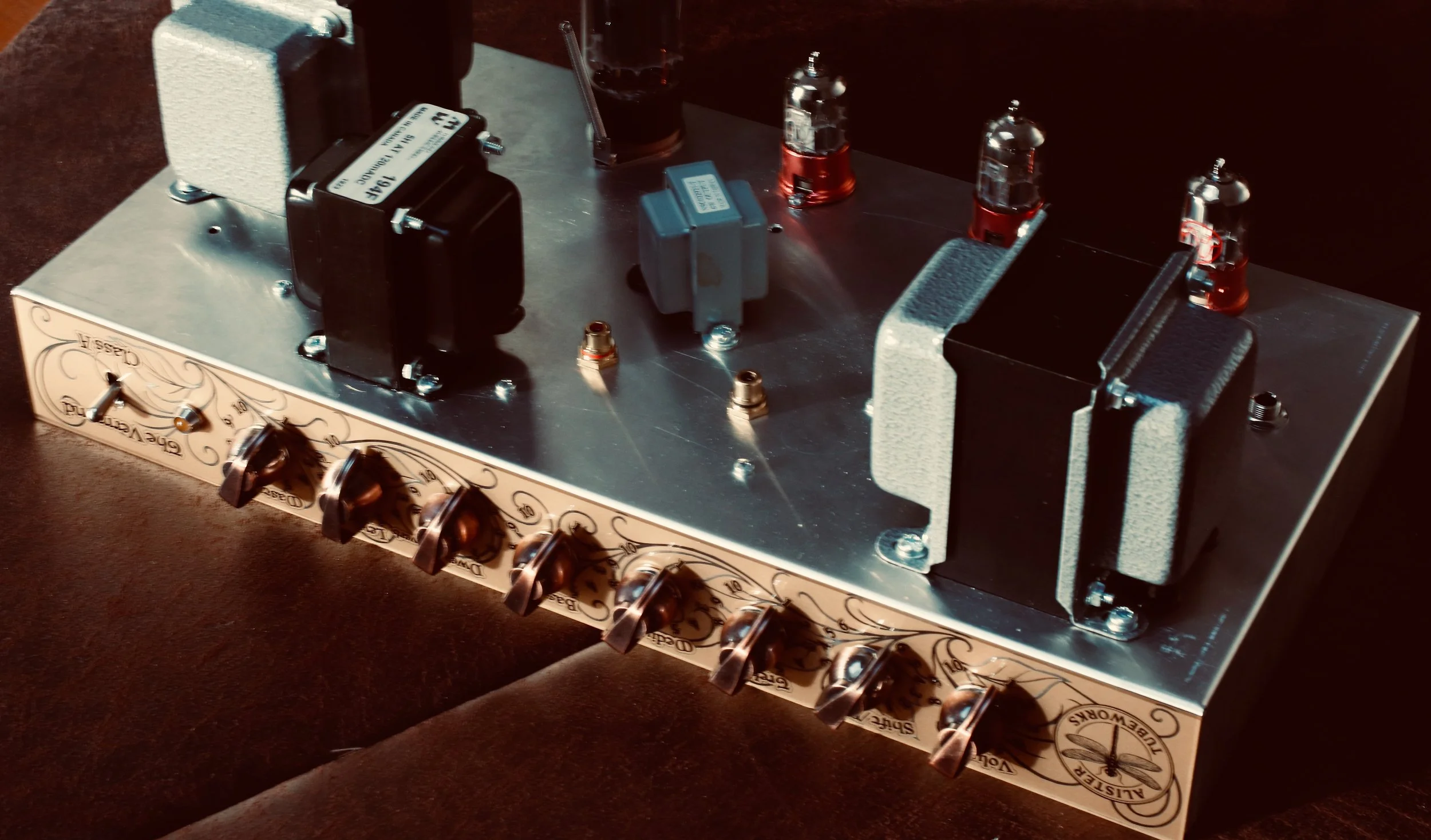 Ampli-tube de guitare électrique avec plusieurs tubes en verre visibles, boutons de contrôle en cuivre à l'avant, monté sur une surface en bois décorée de motifs stylisés.