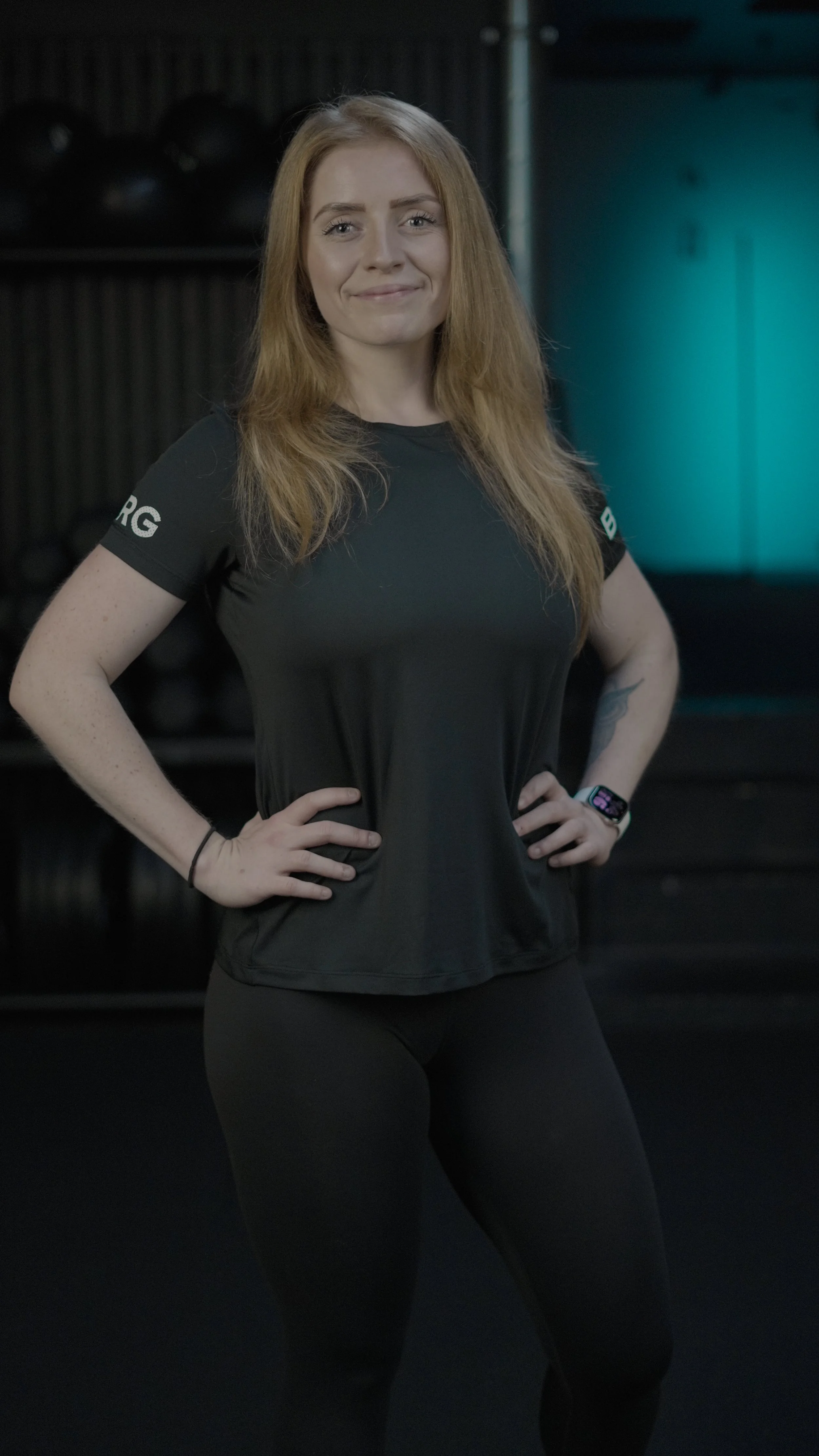 A woman with long, red hair standing in a gym, wearing a black athletic shirt and leggings, with her hands on her hips, smiling.