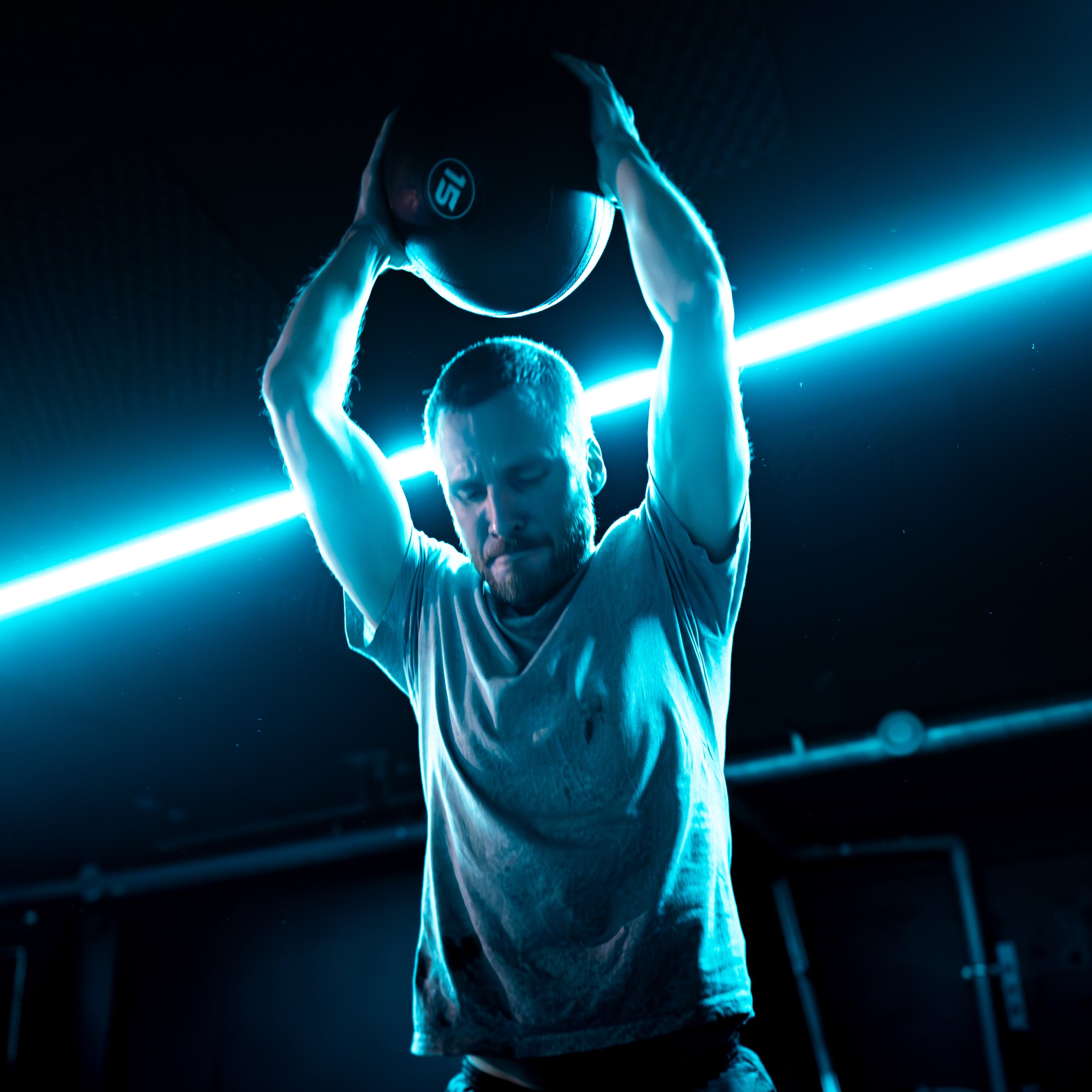 A man lifts a medicine ball overhead in a gym, illuminated by blue lighting, with his eyes closed and a concentrated expression.