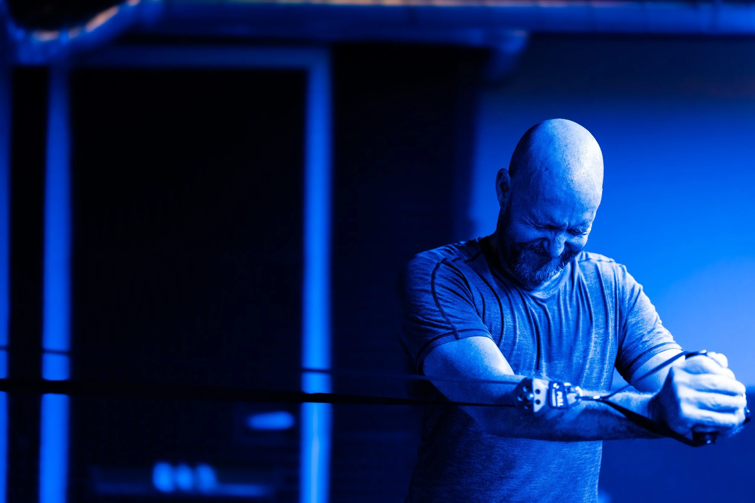 A bald man with a beard, wearing a gray shirt, is smiling and flexing his arm in a blue-lit room, with a dark background.