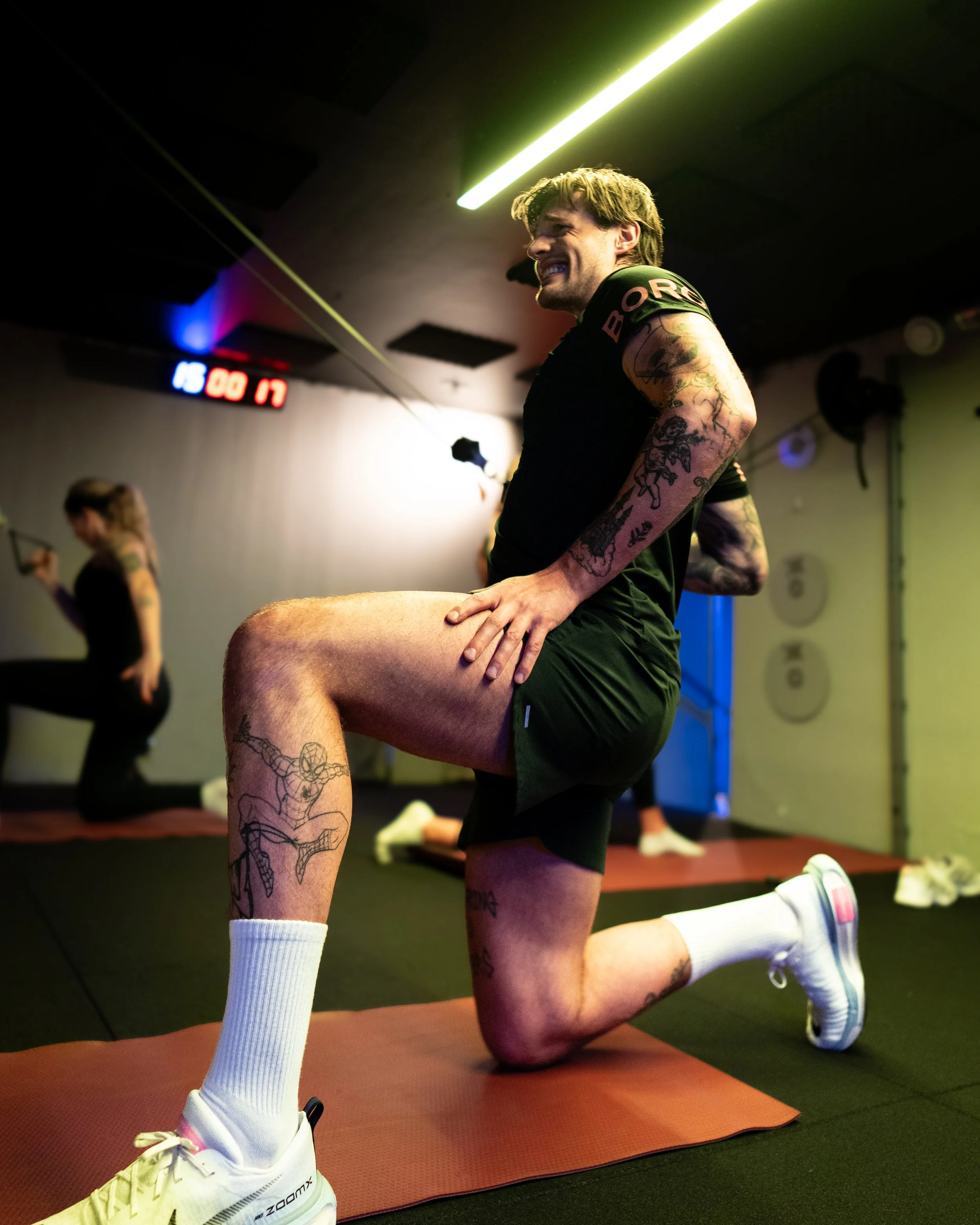 A man with tattoos kneeling on a red workout mat, smiling, in a gym. He is wearing a black t-shirt, black shorts, white socks, and yellow sneakers. There is a woman in the background holding a smartphone, and exercise equipment is visible on the wall.