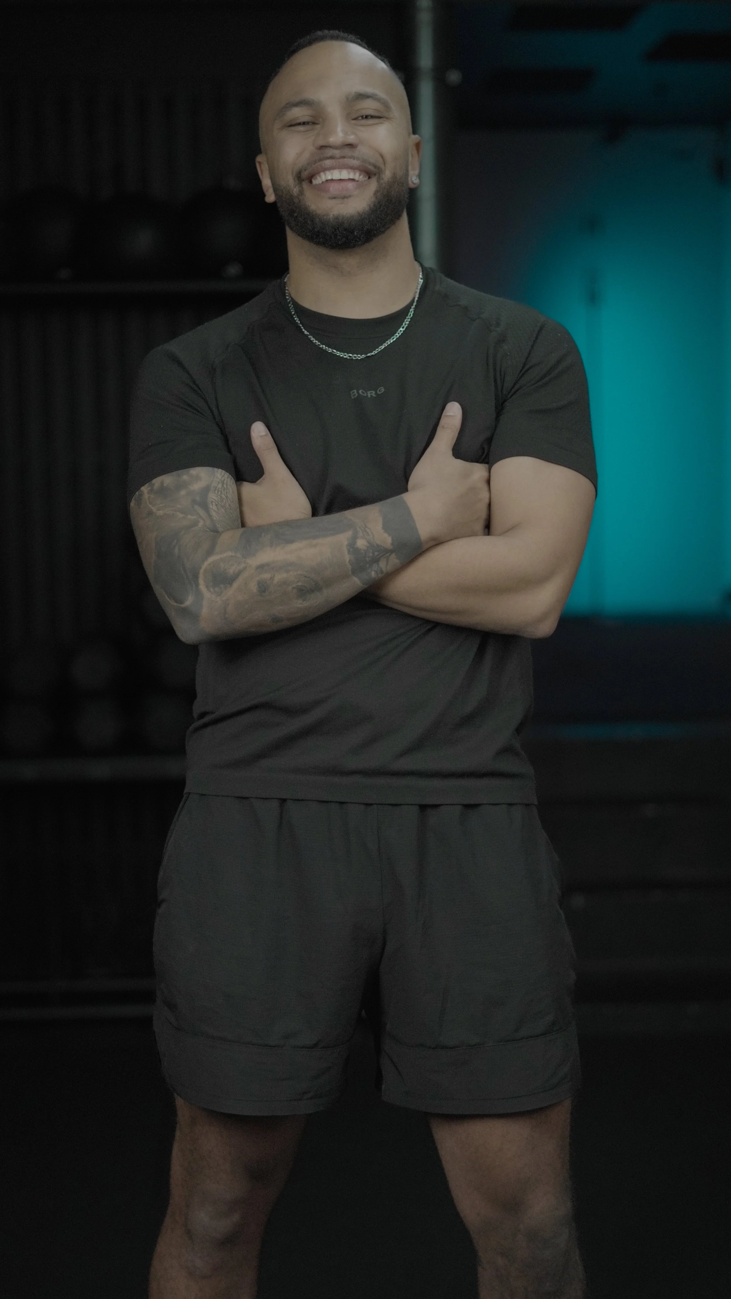 A smiling man with tattoos on his left arm, wearing a black T-shirt and shorts, standing in a gym with gym equipment and black wall padding in the background.