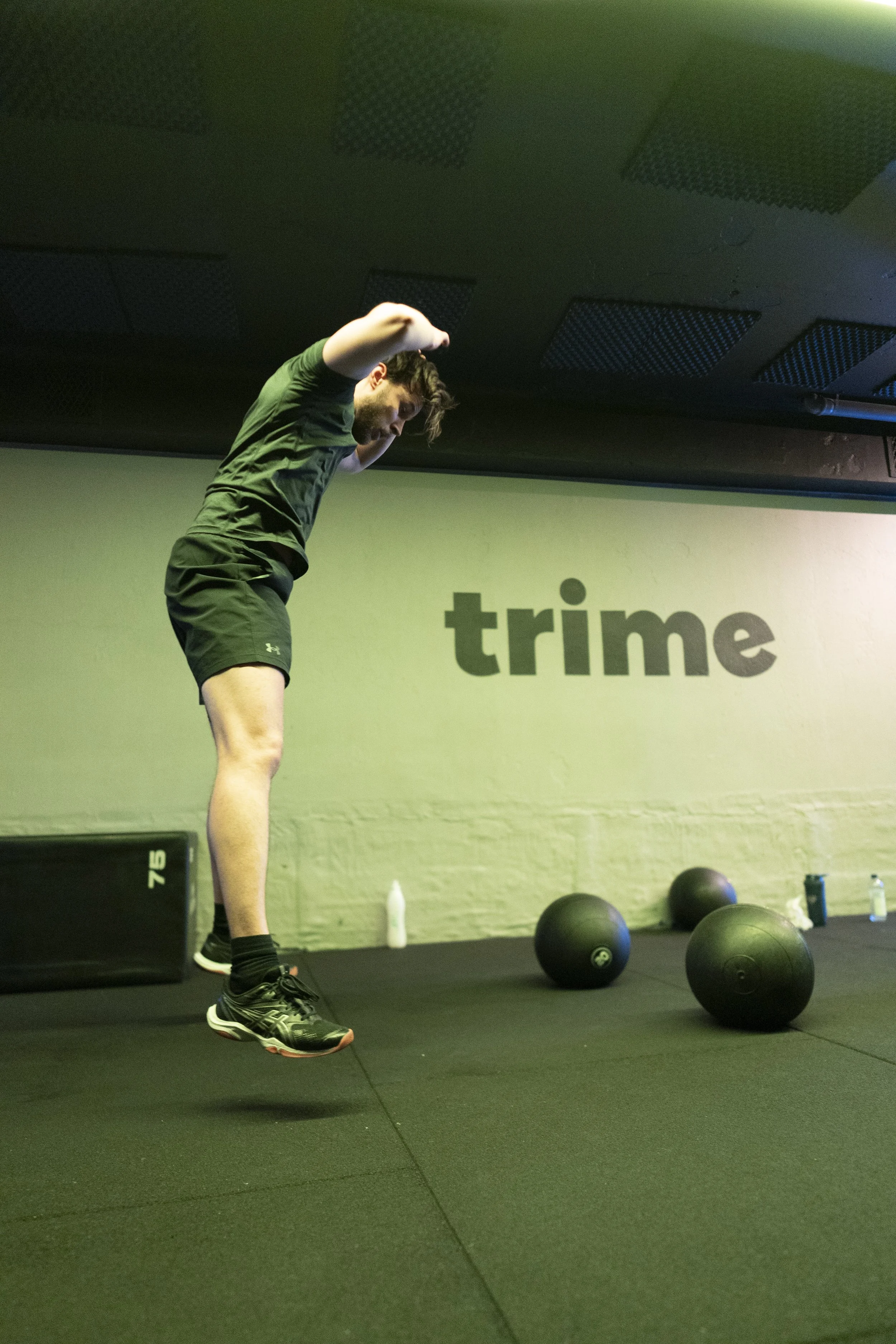 A man jumping in a gym with black medicine balls on the floor and the word 'trime' on the wall behind him.