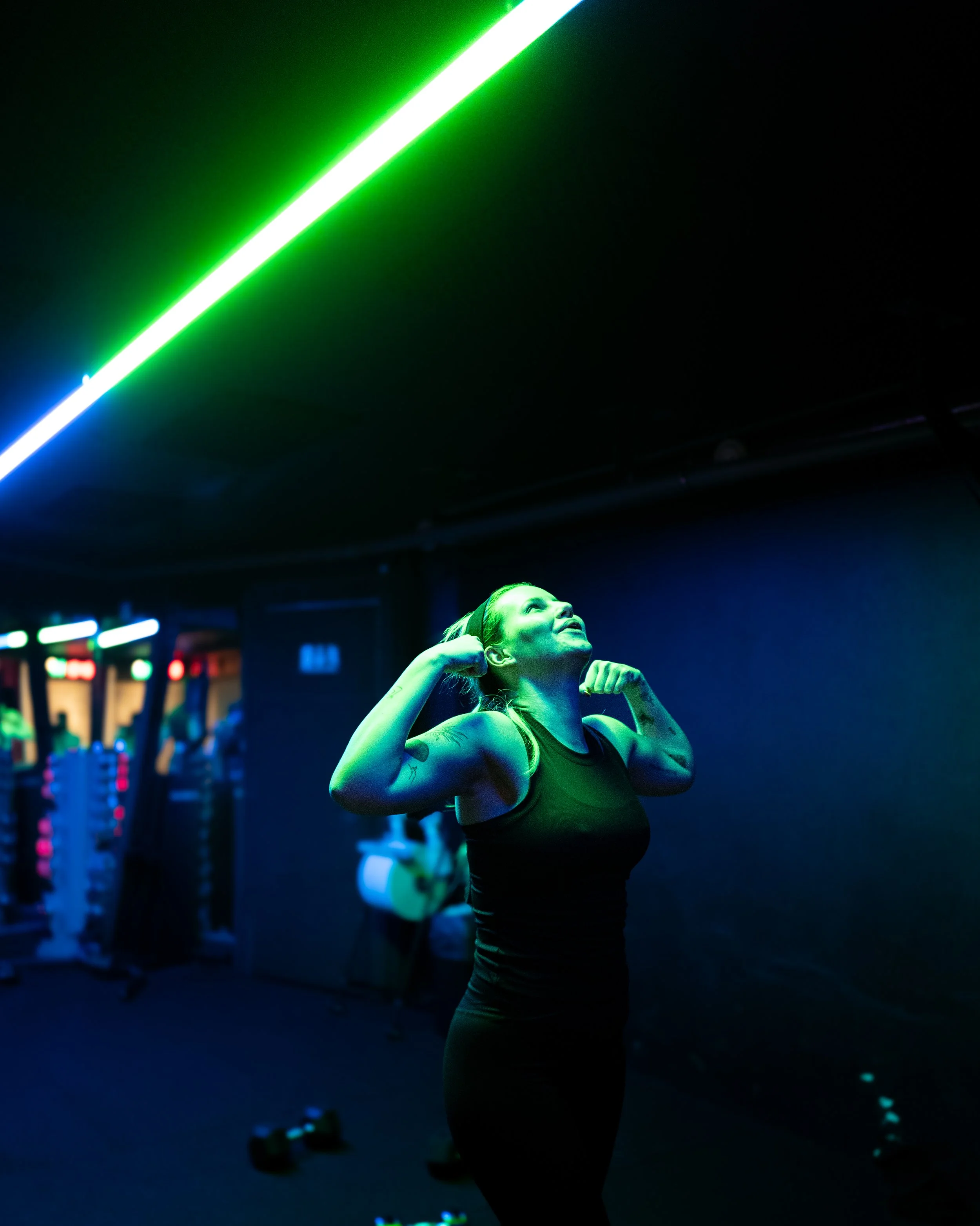 A woman in a workout gym flexing her biceps under a green neon light, with fitness equipment in the background.