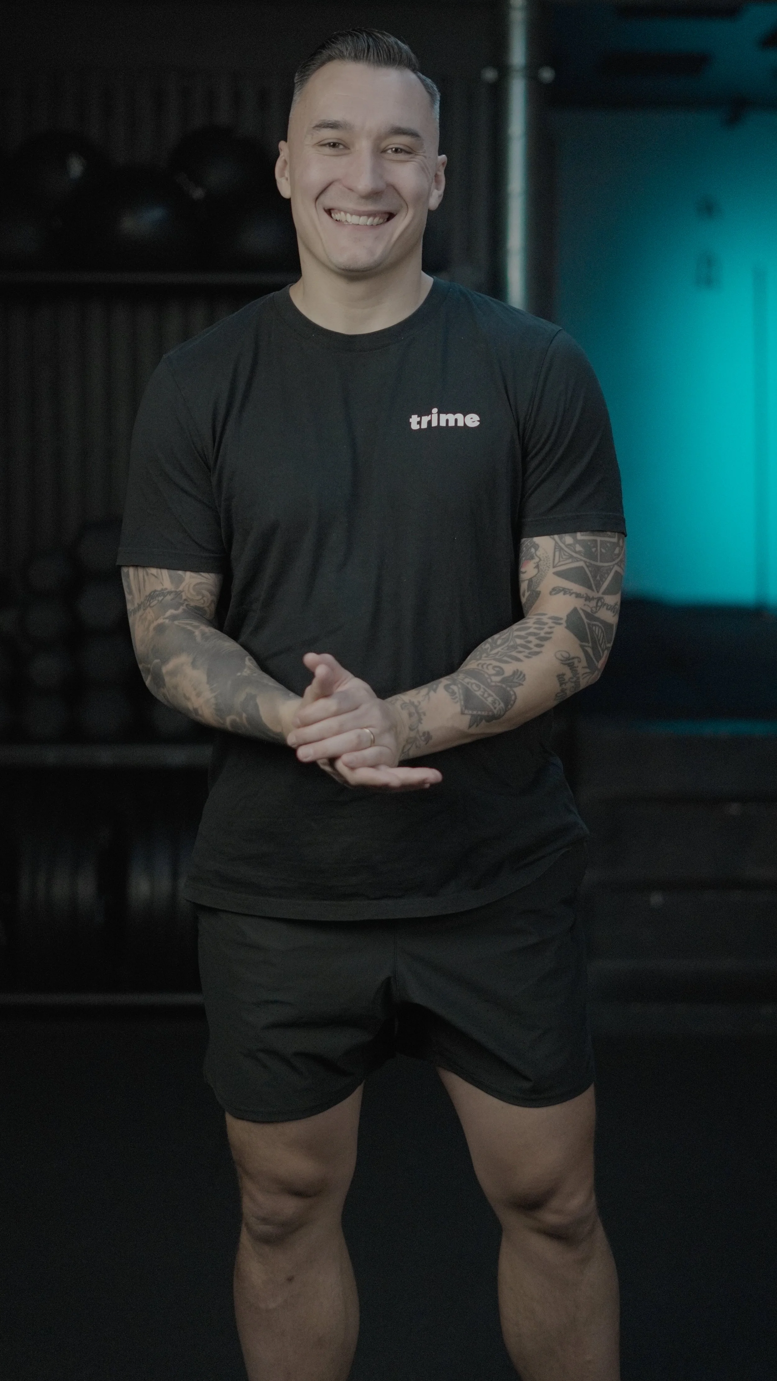 A smiling man in a black t-shirt and black shorts standing in a gym, with muscular legs and tattoos on his arms.