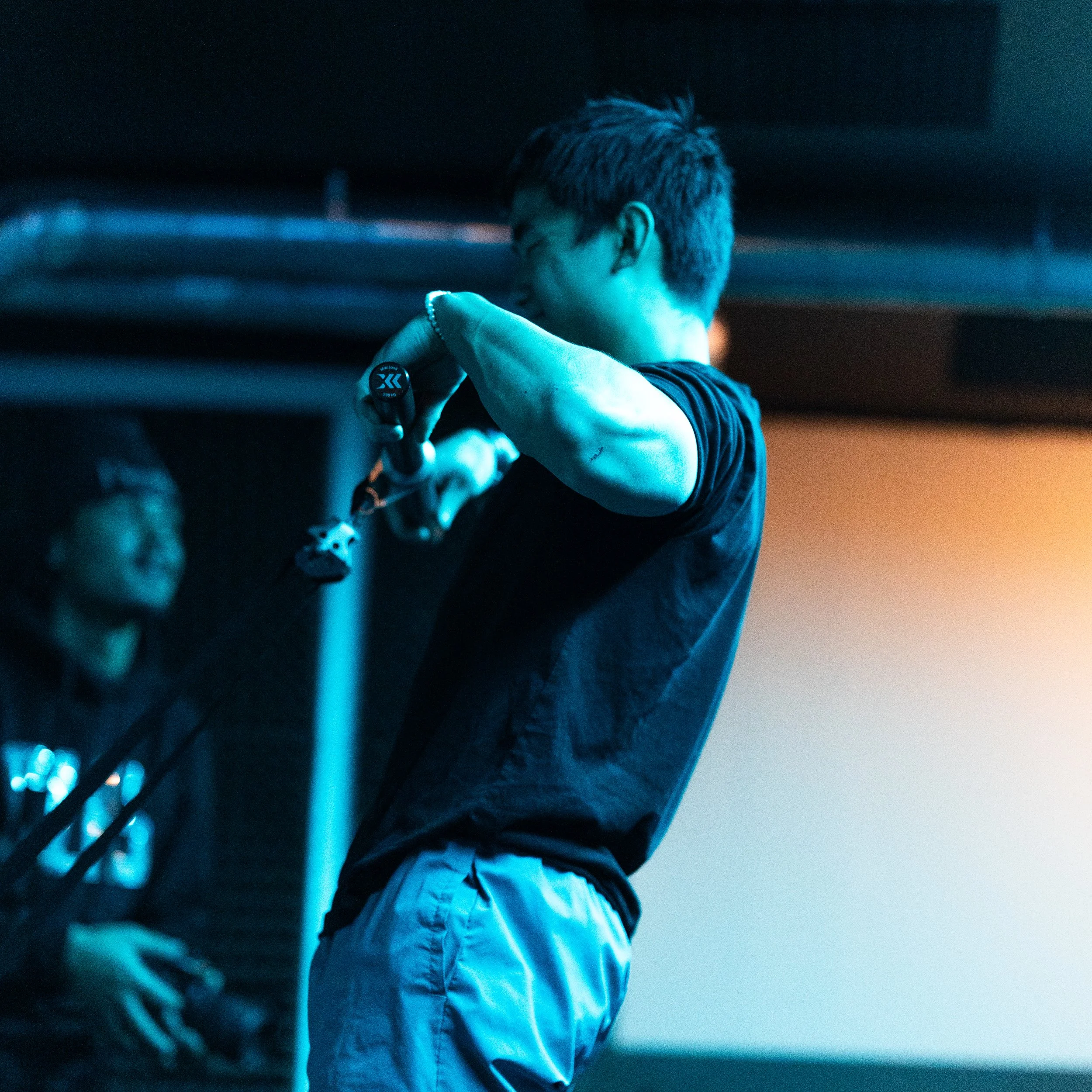 A person with short hair wearing a black shirt and light-colored shorts is holding a small object and looking at it, with a person in the background smiling and wearing glasses.