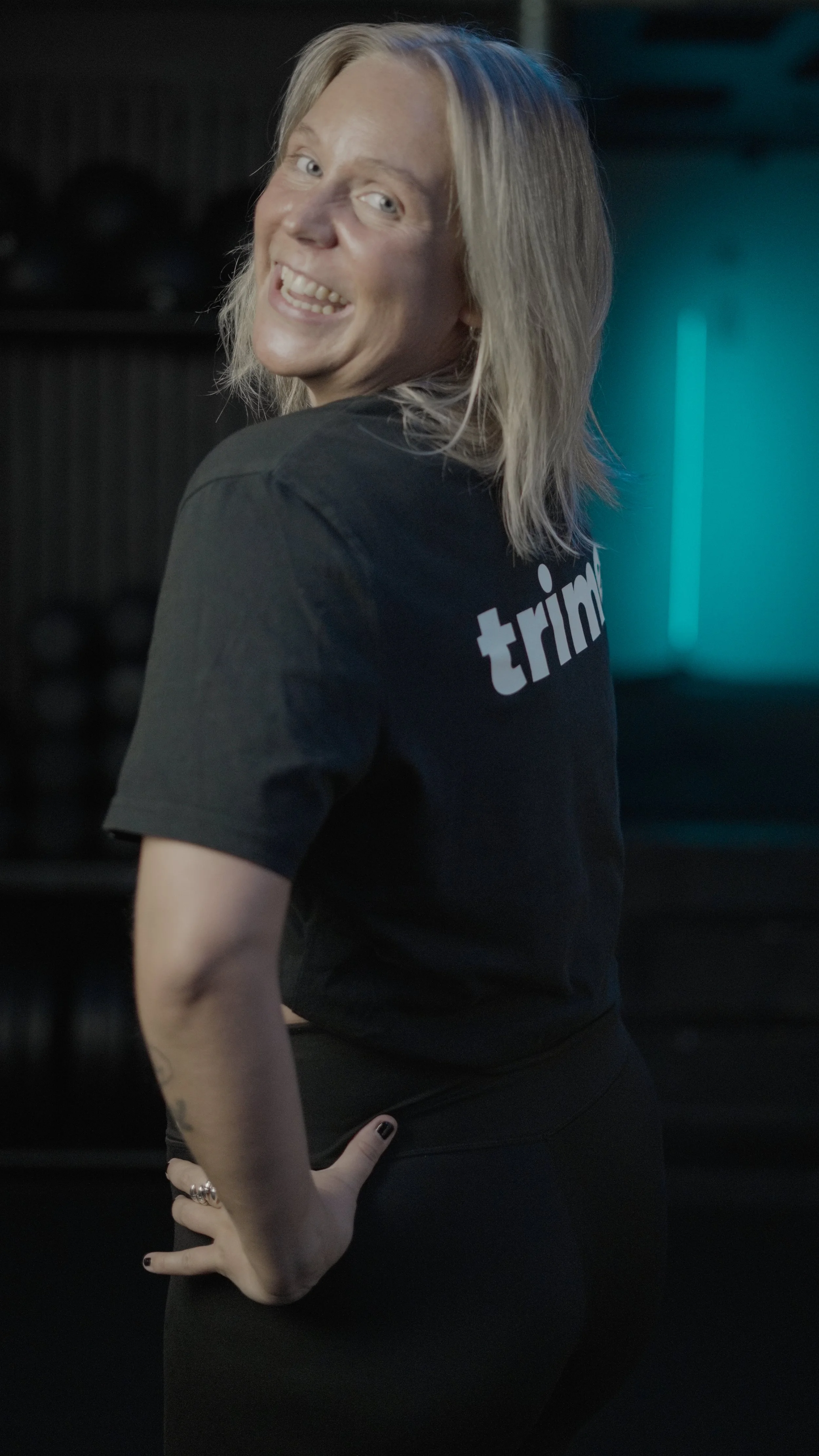A woman with blonde hair smiling, wearing a black T-shirt with partially visible text, standing in a dimly lit room with a blue glow in the background.