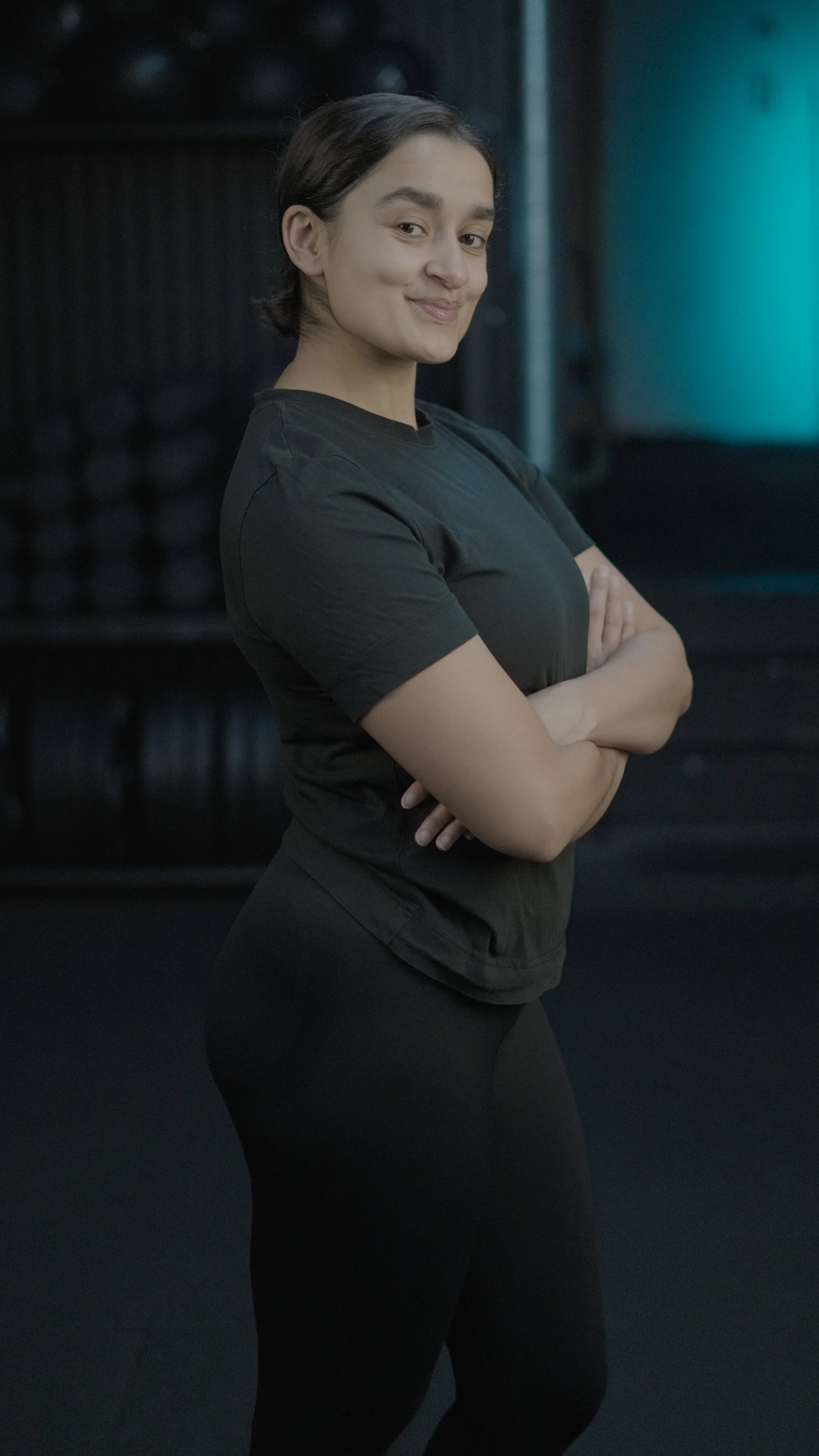 A woman with short dark hair, wearing a black shirt and black leggings, standing with her arms crossed in a gym with blurred weights in the background.