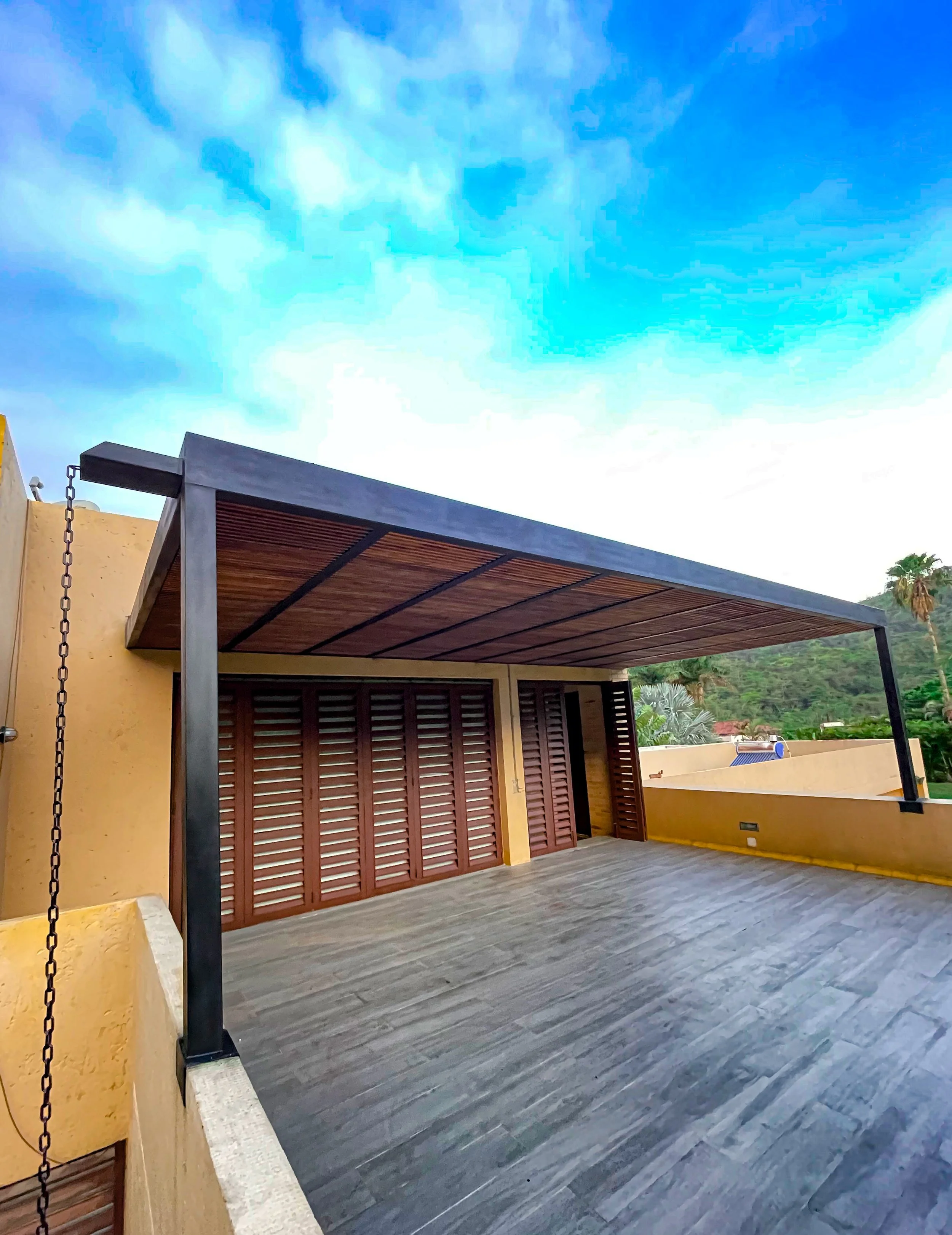 Terraza con pérgola de madera y estructura de metal en un techo de edificio, con paredes de color amarillo y vista a un paisaje con montañas y palmeras.