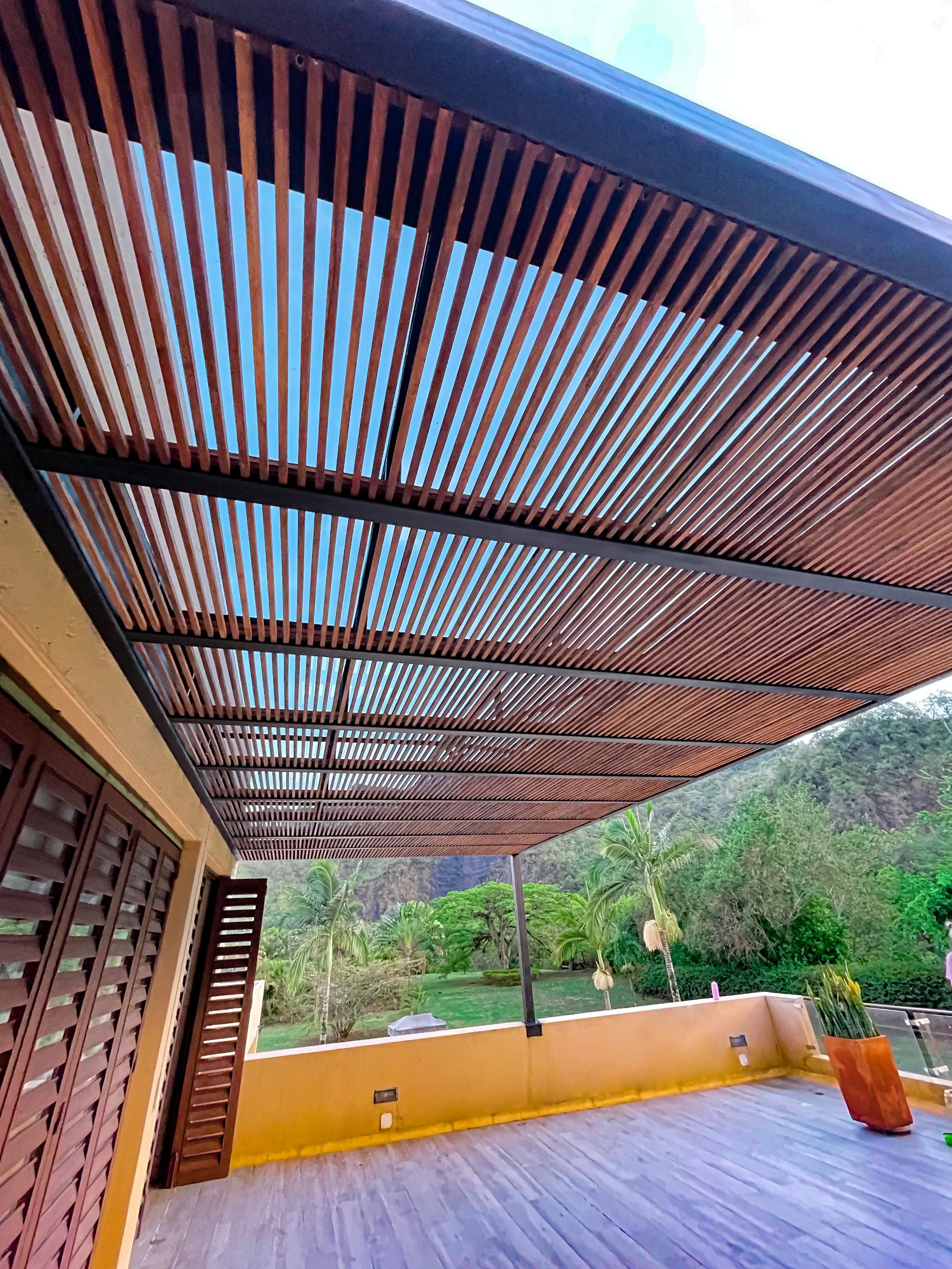 VISTA DE BALCÓN O TERRAZA CON TECHO DE MADERA, CON PLANTA EN MACETA Y VISTAS A ÁRBOLES Y MONTAÑAS AL FONDO.