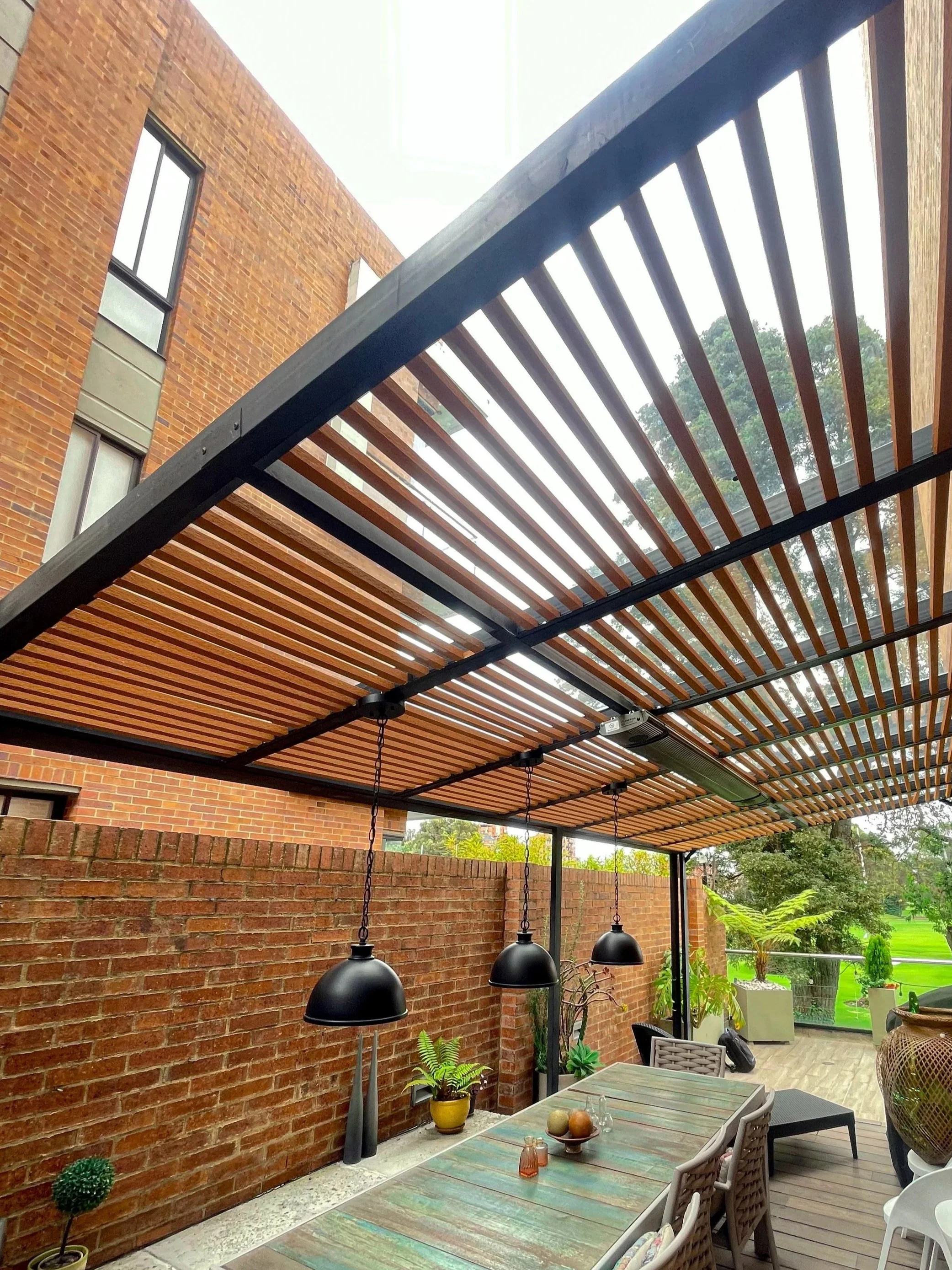 Terraza con toldo de listones de madera, mesas, sillas y plantas en macetas, con vista a un jardín.