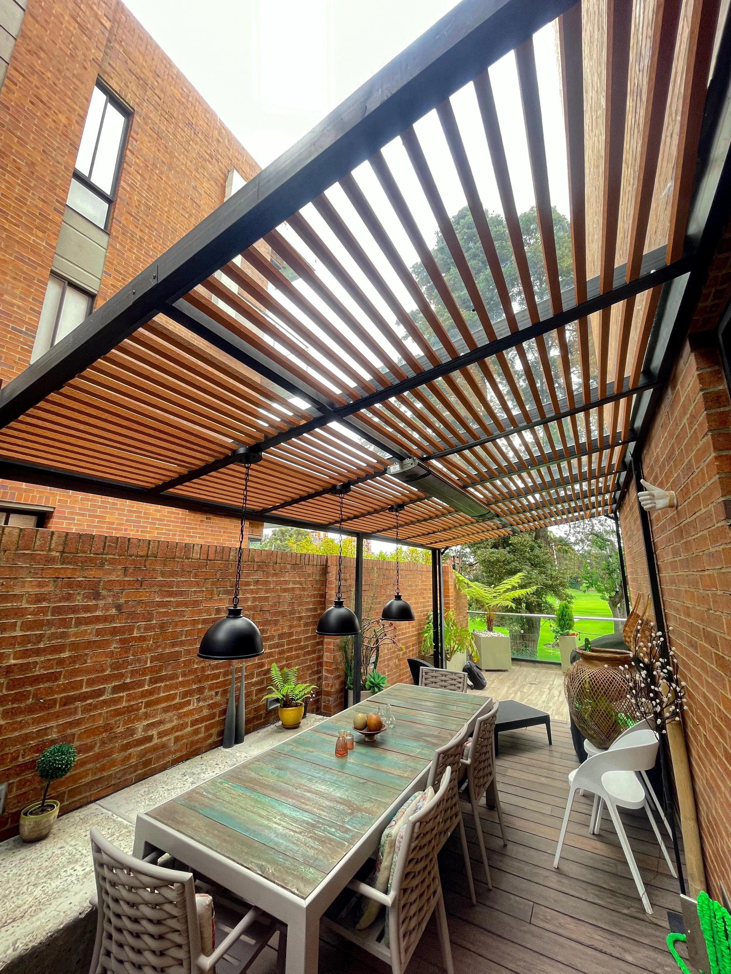 Patio cubierto con techo de listones de madera y lámparas negras colgantes, mesa de comedor con sillas, plantas decorativas y vista al jardín con árboles verdes.