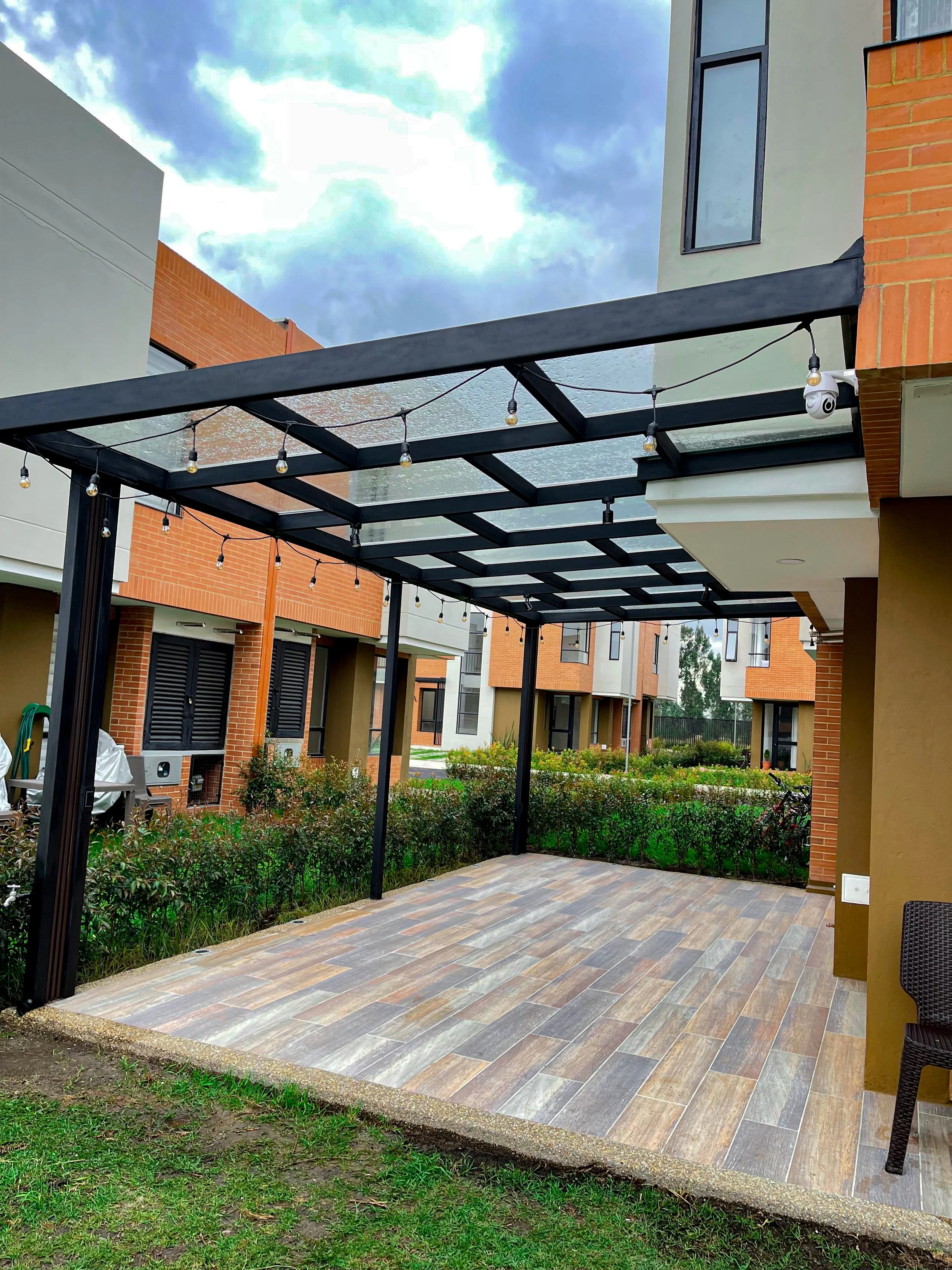 Patio exterior con estructura de pergola negra con luces decorativas. Piso de cerámica en tonos marrones y verdes. Al fondo, edificios residenciales con paredes de ladrillo y ventanas. Áreas verdes con arbustos y césped.