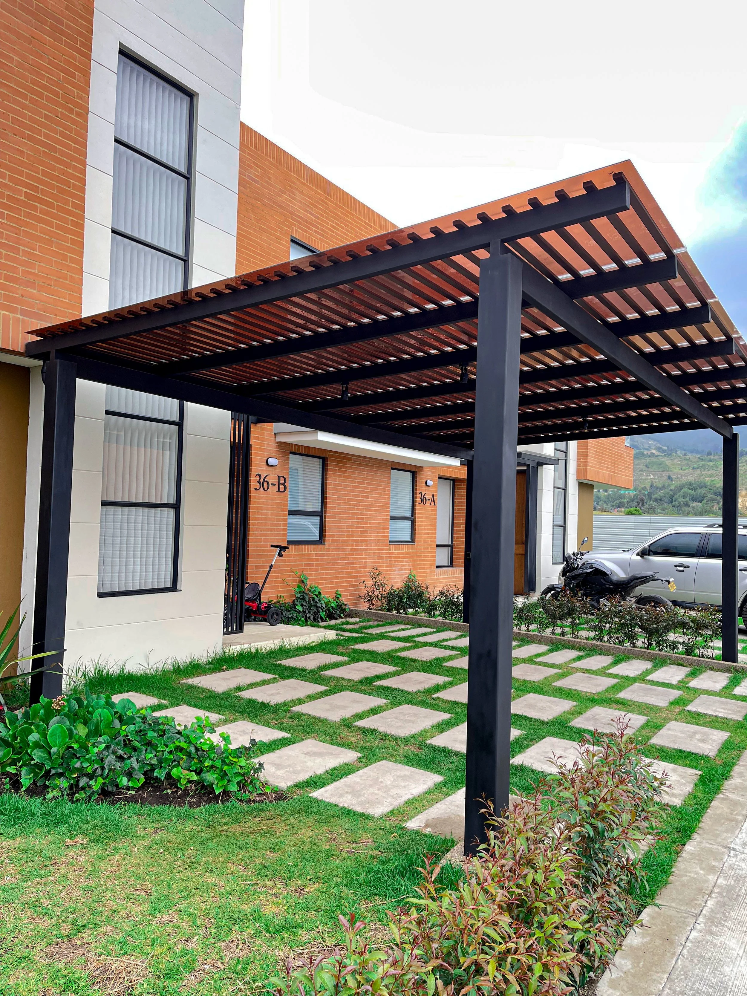 Carport con estructura de columnas negras y techo de listones de madera, en zona de jardín con césped y macetas. Edificio de apartamentos de ladrillo y ventanas con persianas.