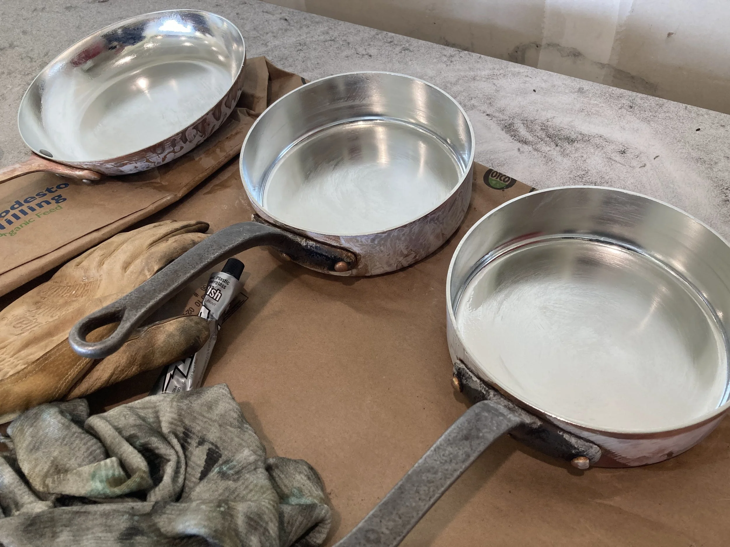 Three freshly tinned copper pots, a marker, a pair of gloves, and a pair of worn leather gloves on a work surface.
