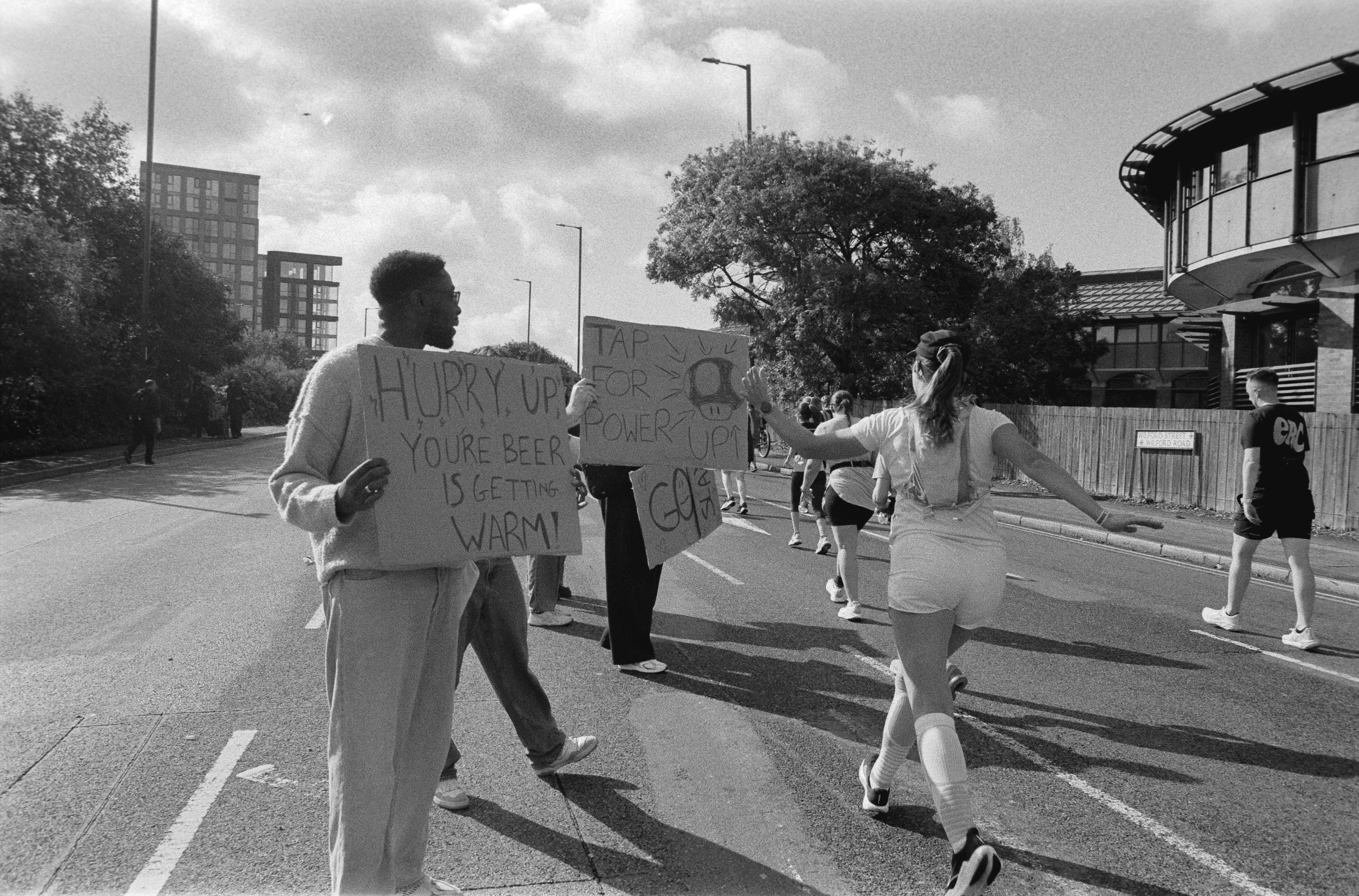 People participating in a street protest or march, holding signs with messages about climate change and electricity, with some making gestures, on a city street with trees and buildings in the background.