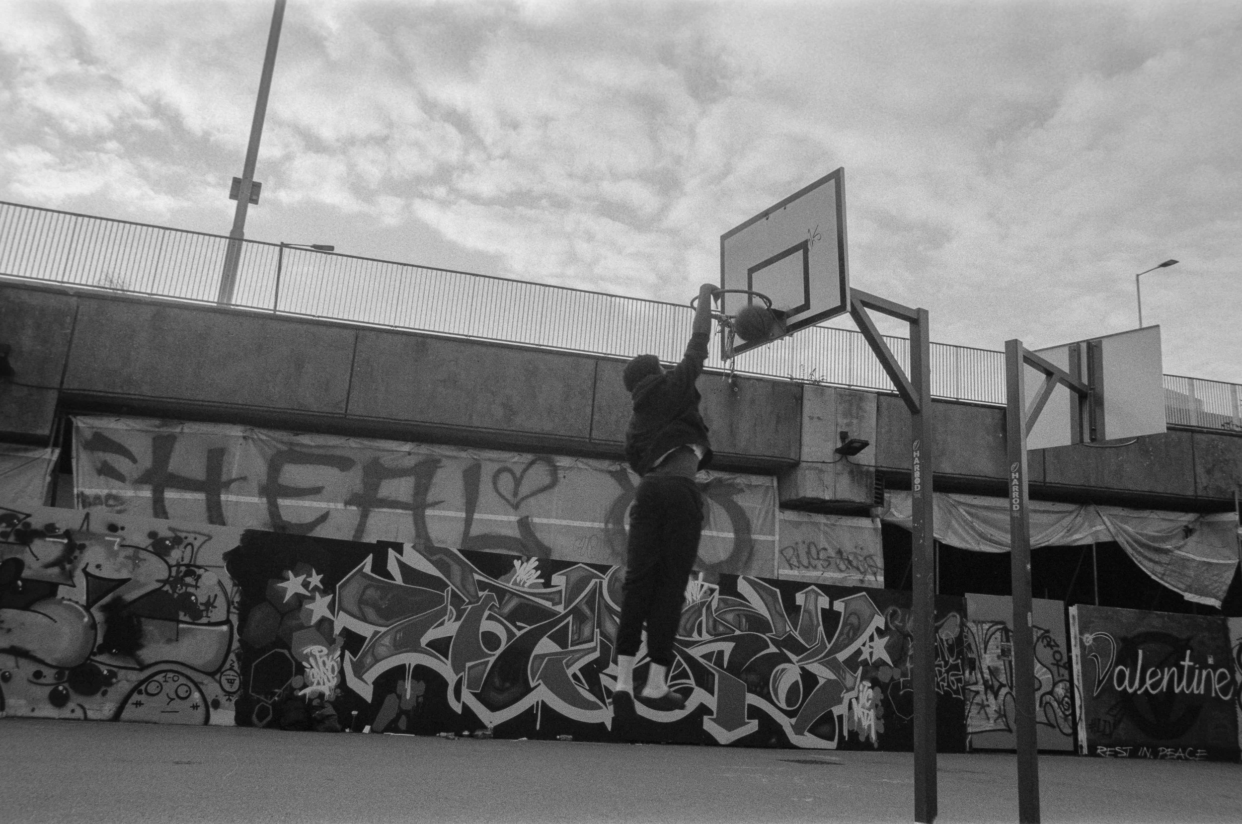 A person jumping to dunk a basketball in an outdoor court with graffiti on the wall and a cloudy sky.