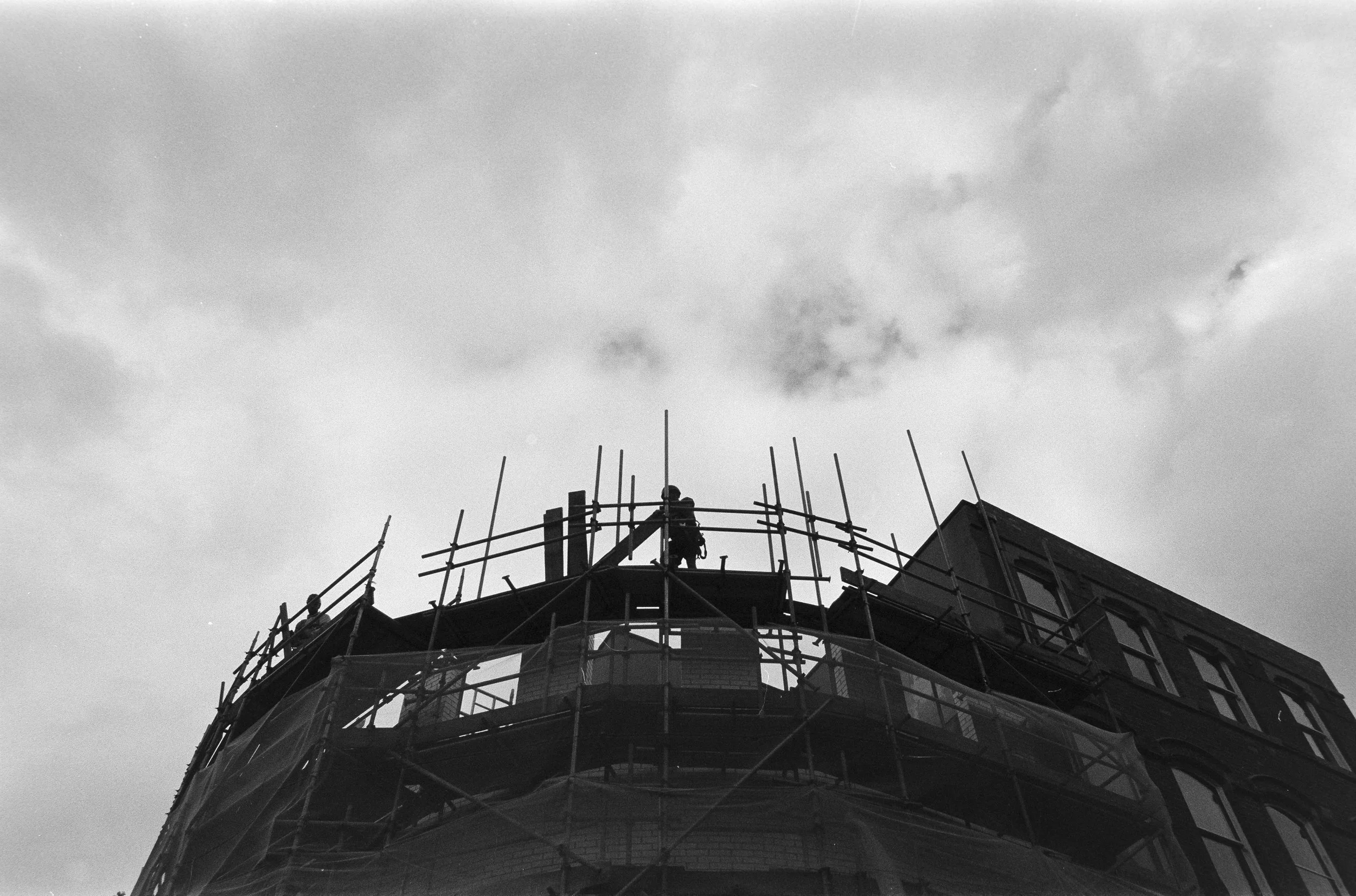 Black and white photo of a building under construction or renovation with scaffolding around the top, and a worker standing on the scaffolding near the center of the image. The sky is cloudy in the background.