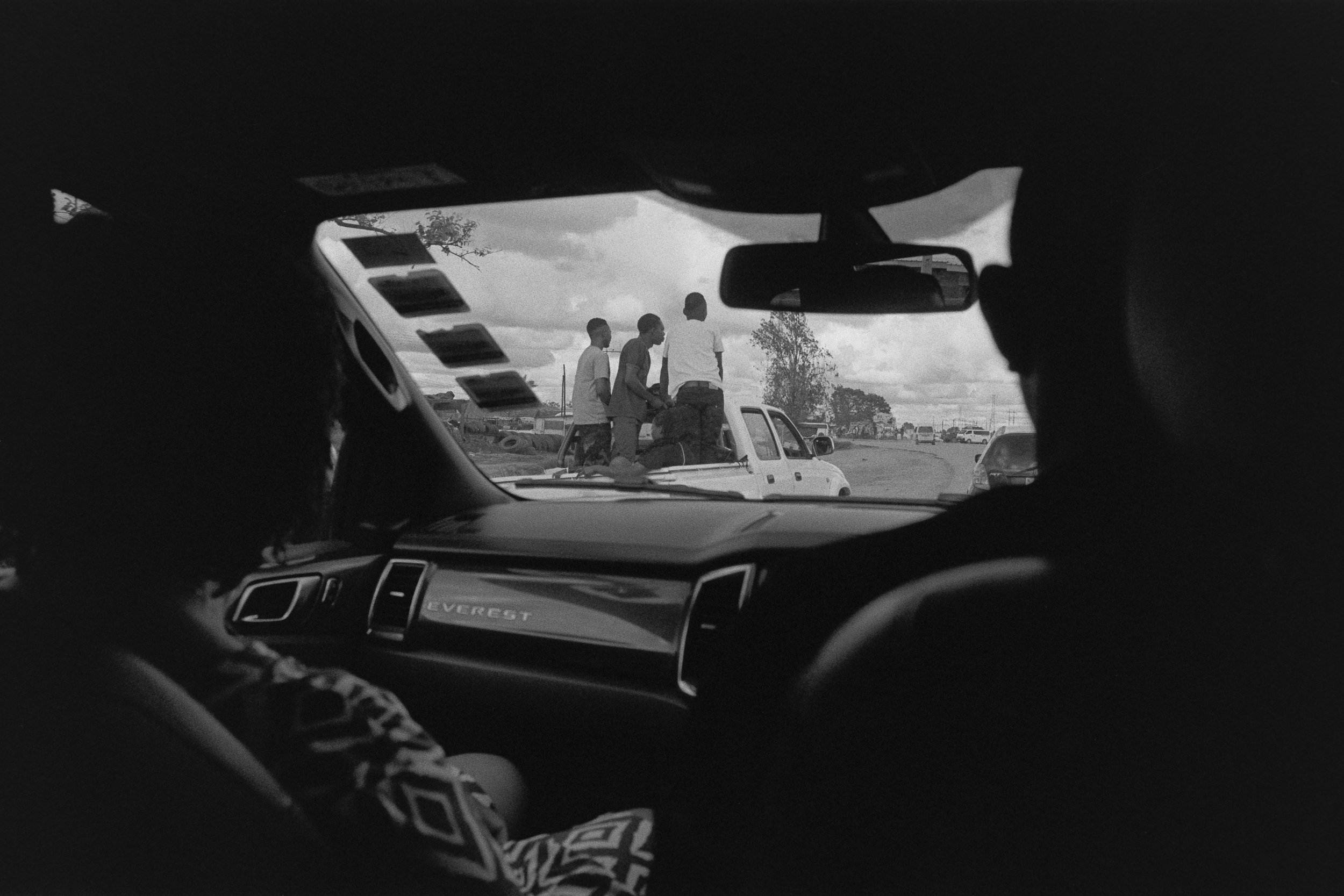 A black and white photo taken from inside a vehicle showing four people sitting on the back of a pickup truck, visible through the windshield, with cars and trees in the background.