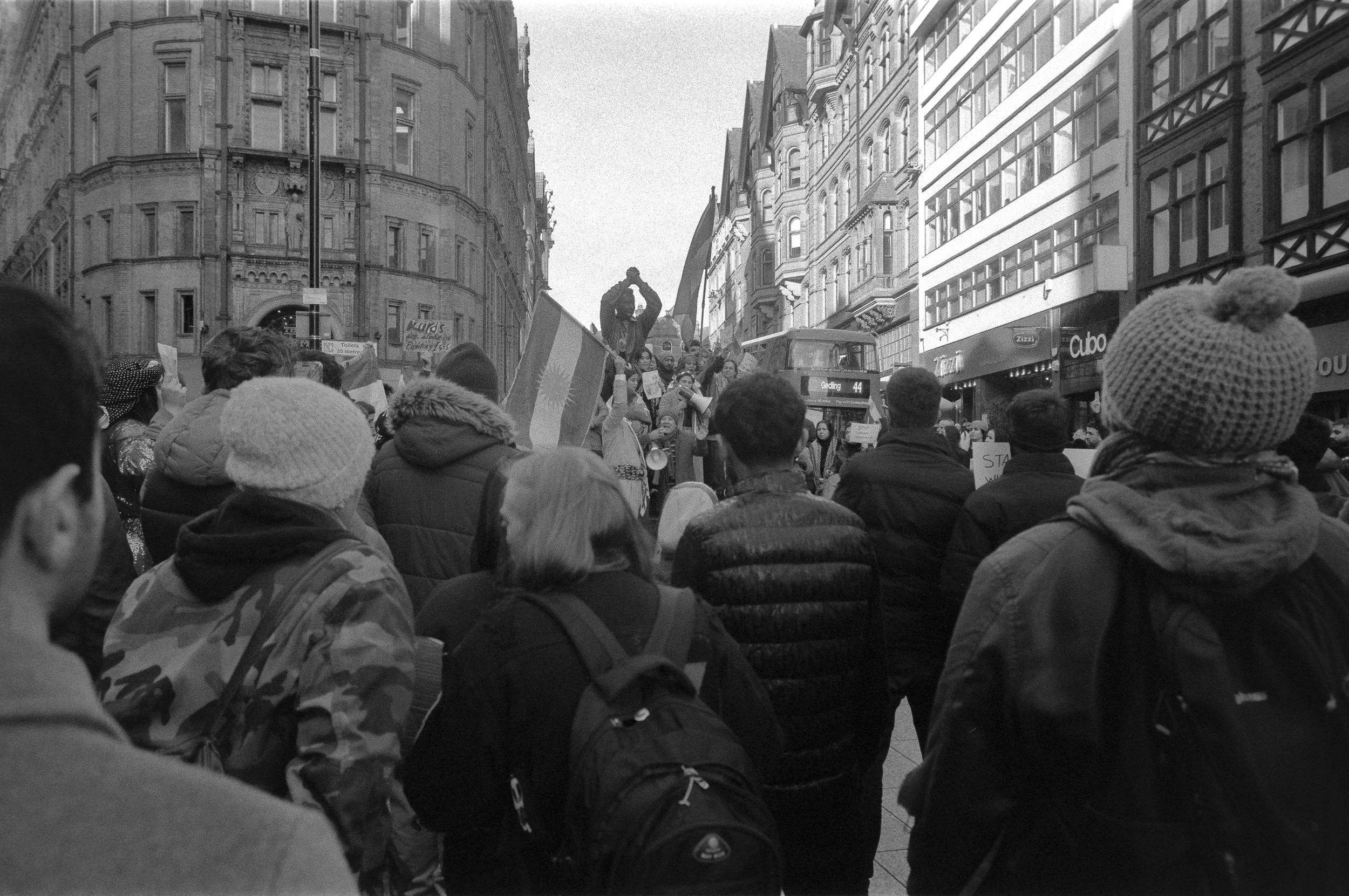Crowd of people gathered on city street, protesting or demonstrating, holding flags and signs, with buildings and a double-decker bus in the background.