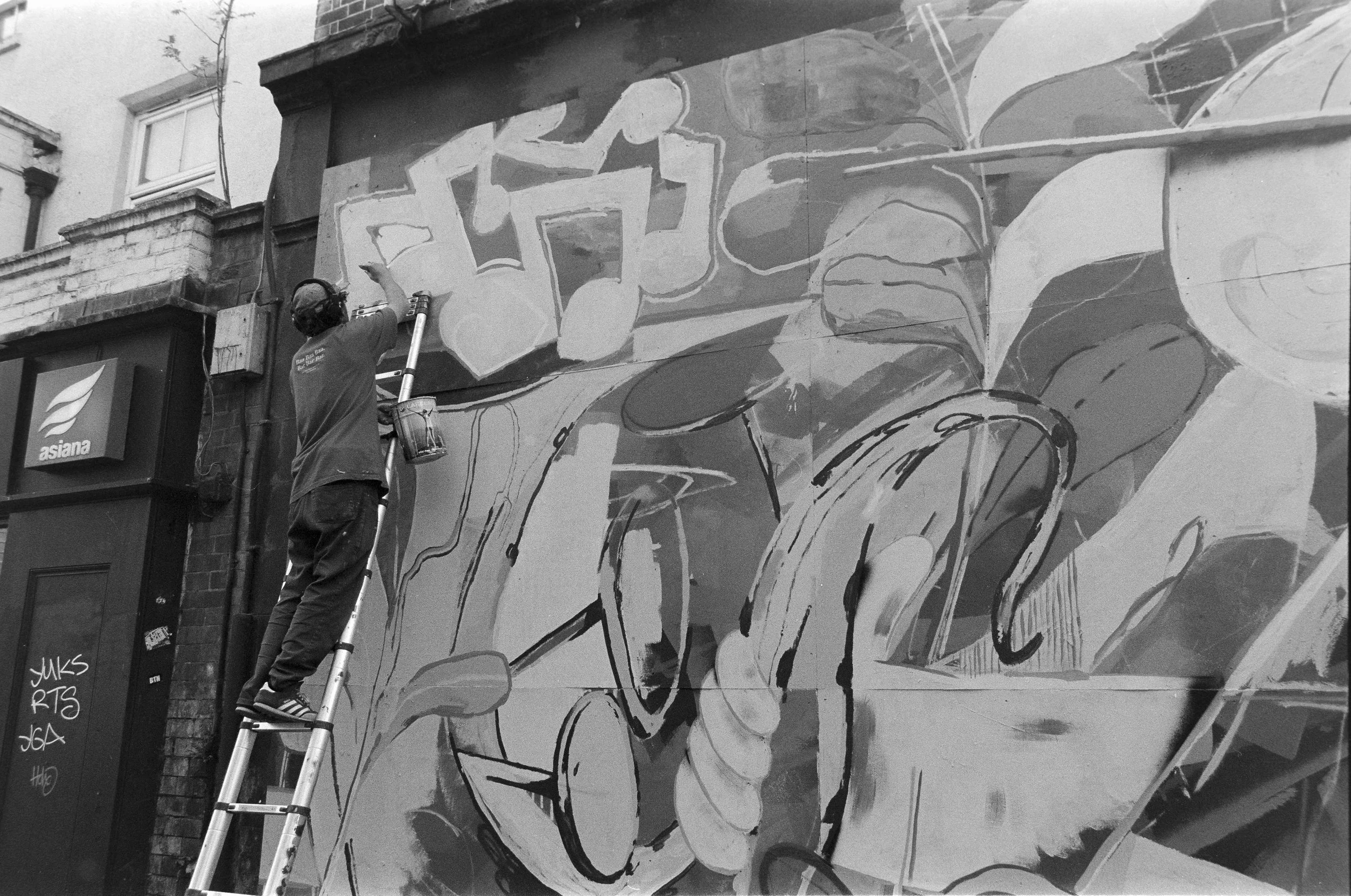A person wearing headphones is working on a large abstract mural on a building wall, using a ladder to reach higher sections.