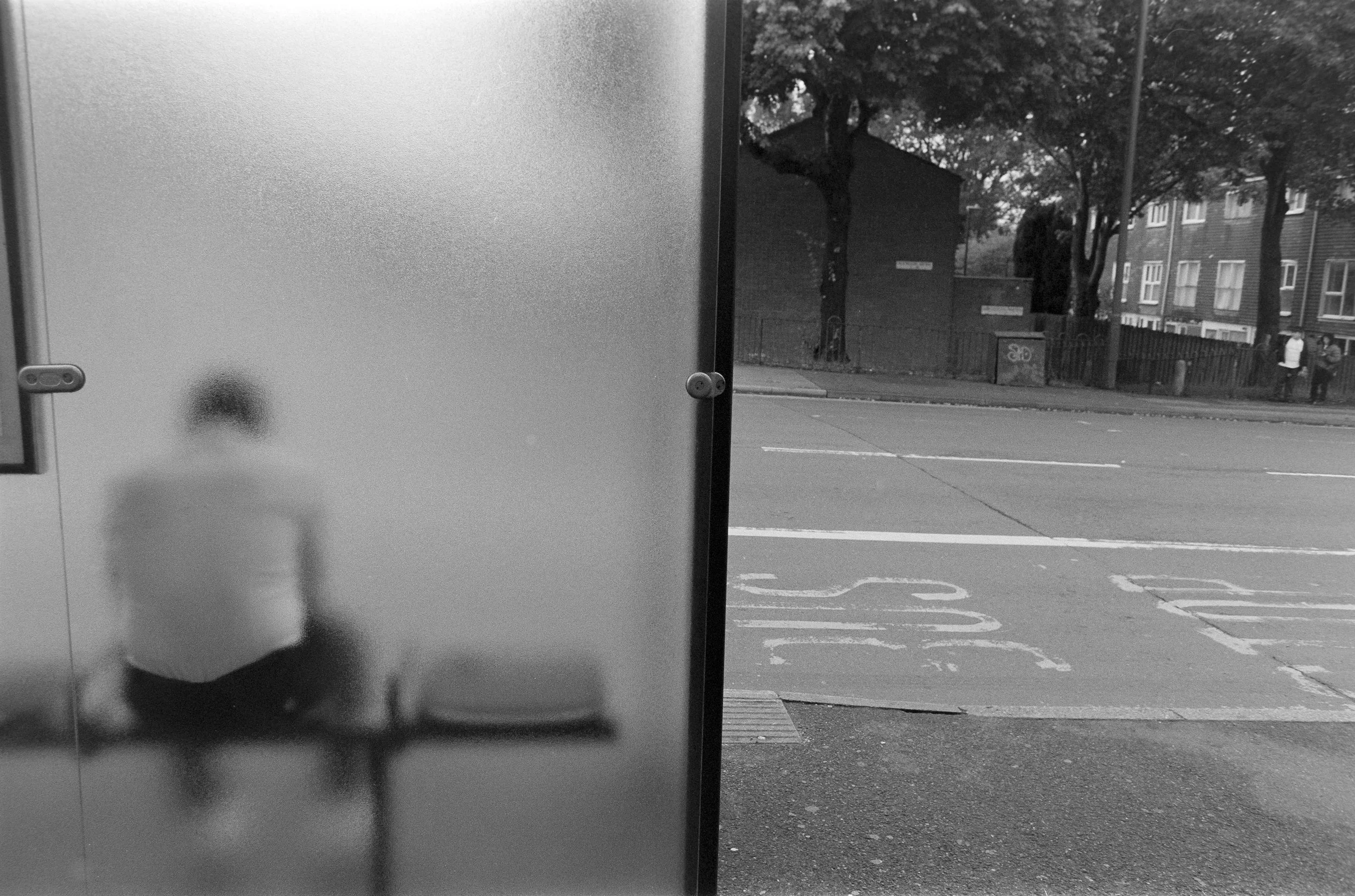 A person sitting in front of a frosted glass panel, seen through the blurred glass, with an outdoor street scene including trees, buildings, and a few people in the background.