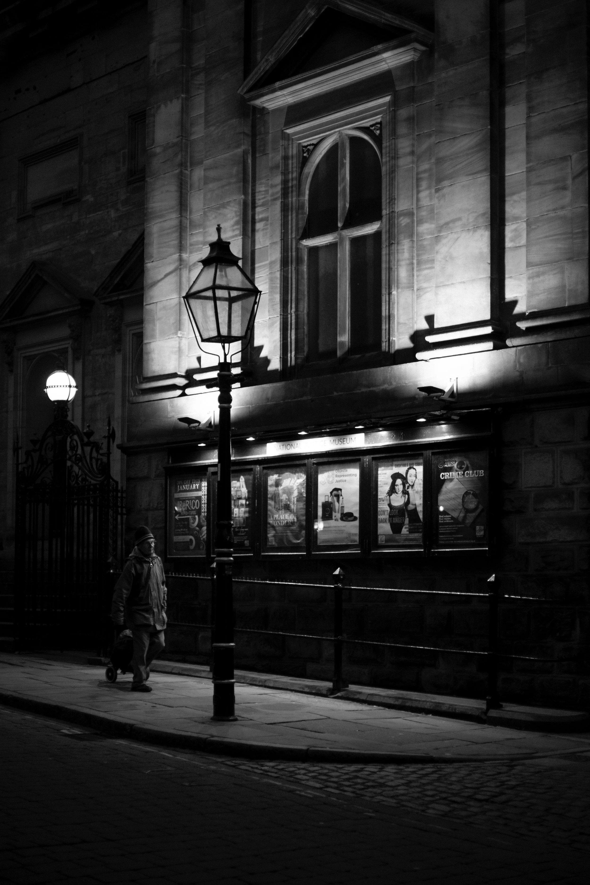 A person walking with a rolling suitcase on a city sidewalk at night. The scene is illuminated by street lamps and an adjacent building with posters in a display case. The building has large arched windows and detailed stone architecture.