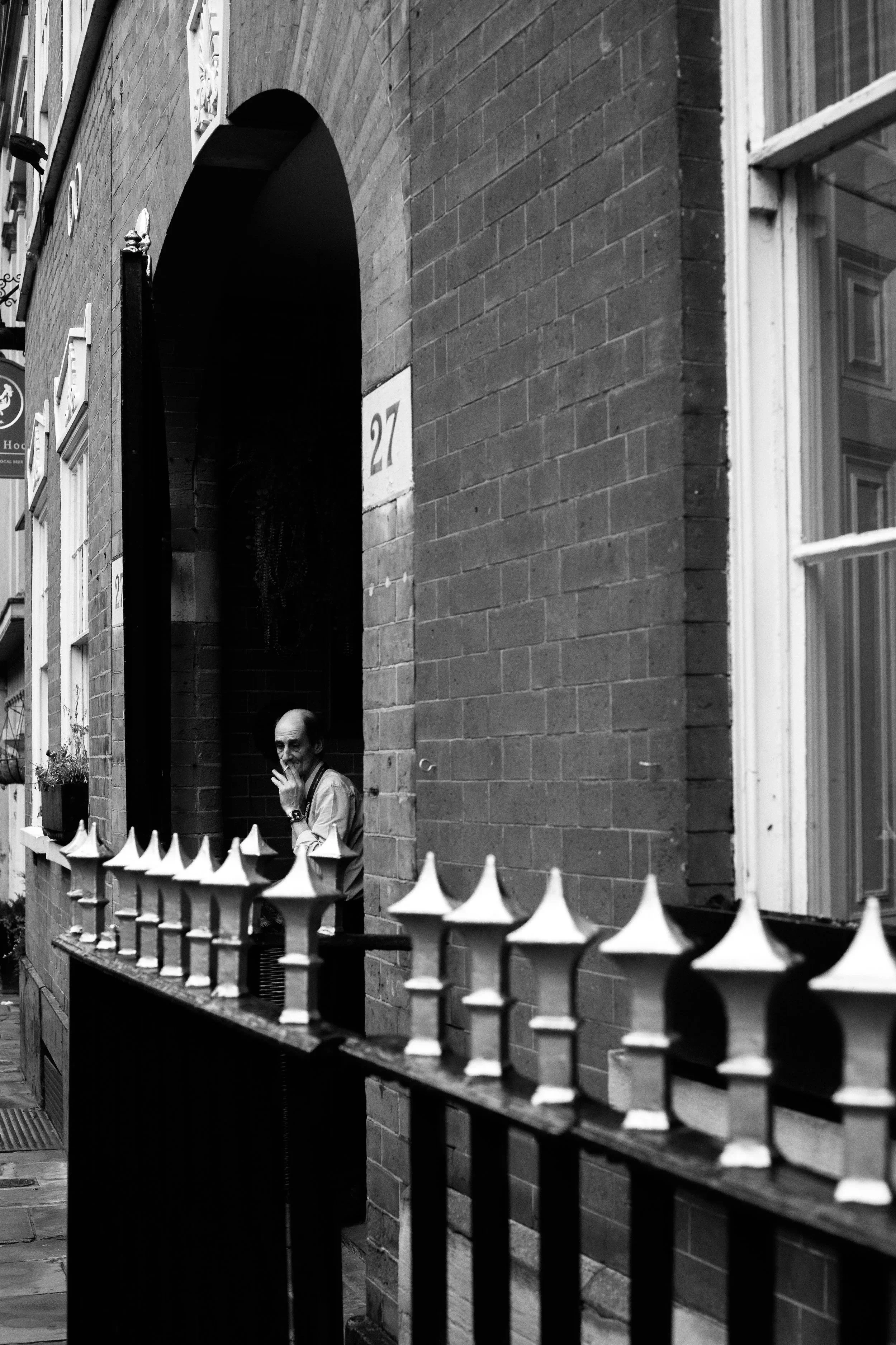 A man standing on a sidewalk behind a decorative metal fence, looking through a window of a brick building with the number 27, near an arched doorway.