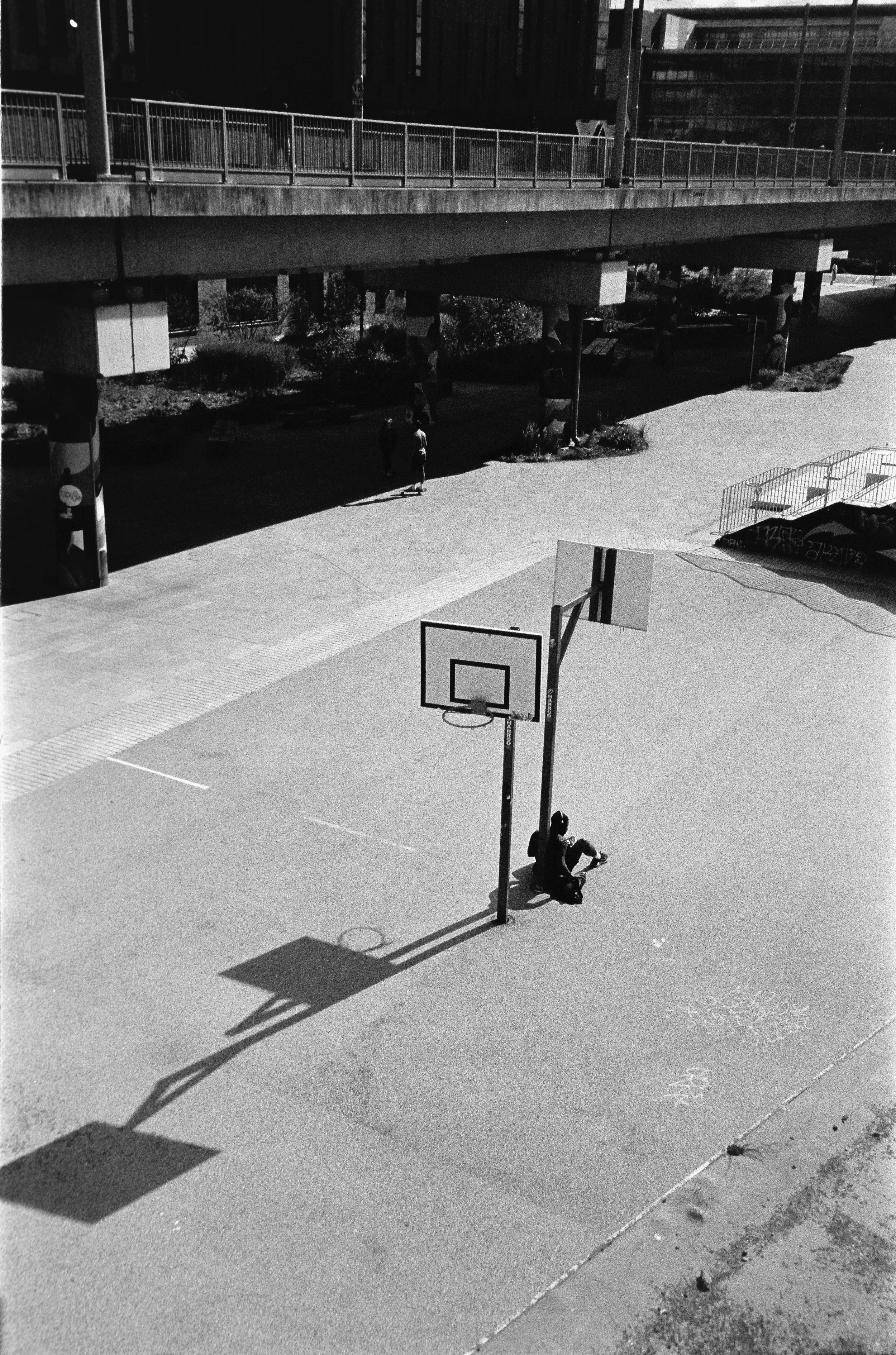 A black and white photo of an outdoor basketball court with a single person sitting under the hoop and two more walking underneath a bridge in the background.