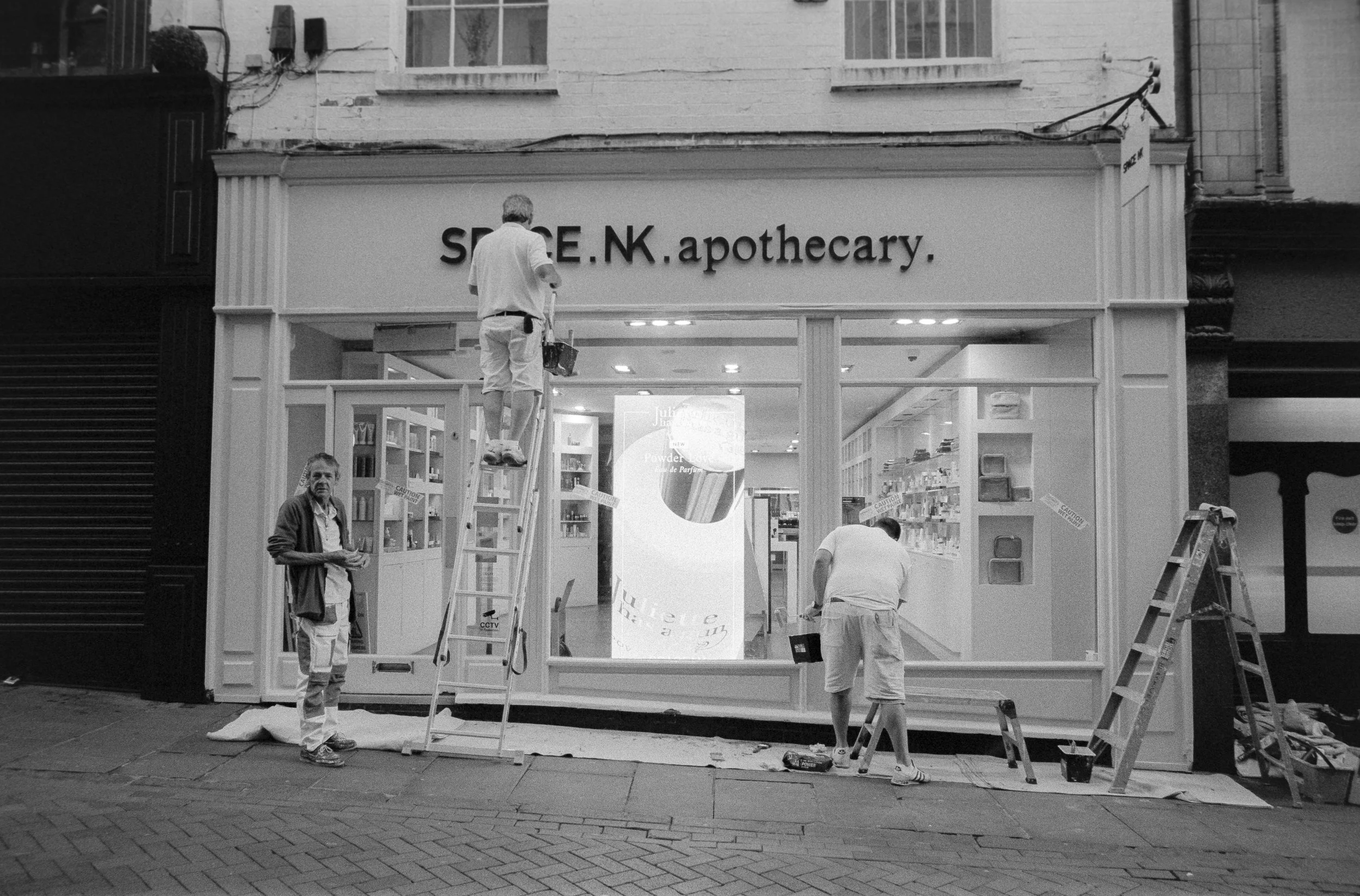 Workers installing or repairing the storefront of a pharmacy called 'SPACE.NK.apothecary' on a city street. One worker is on a ladder, another stands outside holding tools, and another is working along the bottom of the storefront window. The store h
