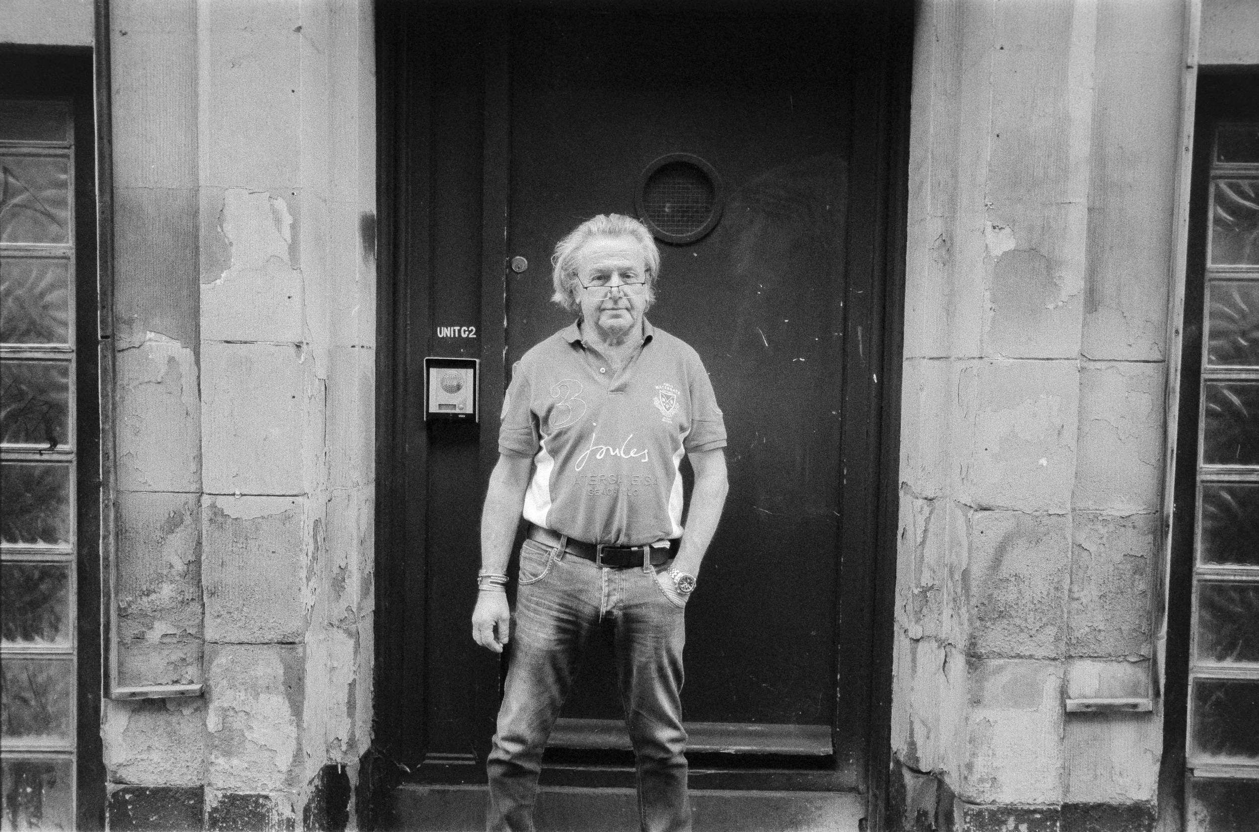 A man with long, curly hair and glasses standing outside a door with his hand in his pocket. He is wearing a printed polo shirt and jeans. The door behind him is black with a small circular window and a keypad entry system, surrounded by worn brick a