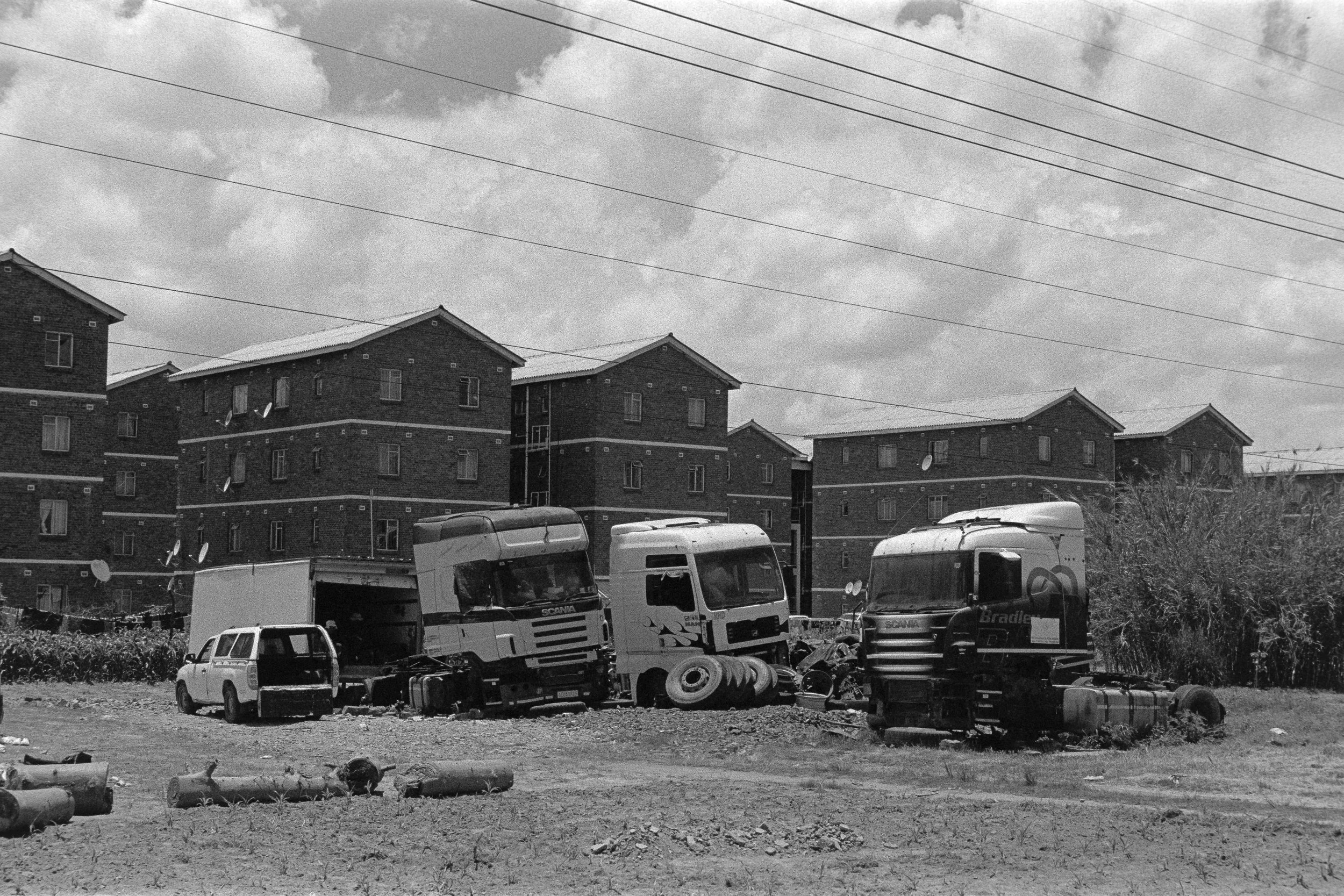 A black and white photo of a junkyard with several abandoned and broken-down trucks and a small car, with residential brick buildings in the background and a cloudy sky above.