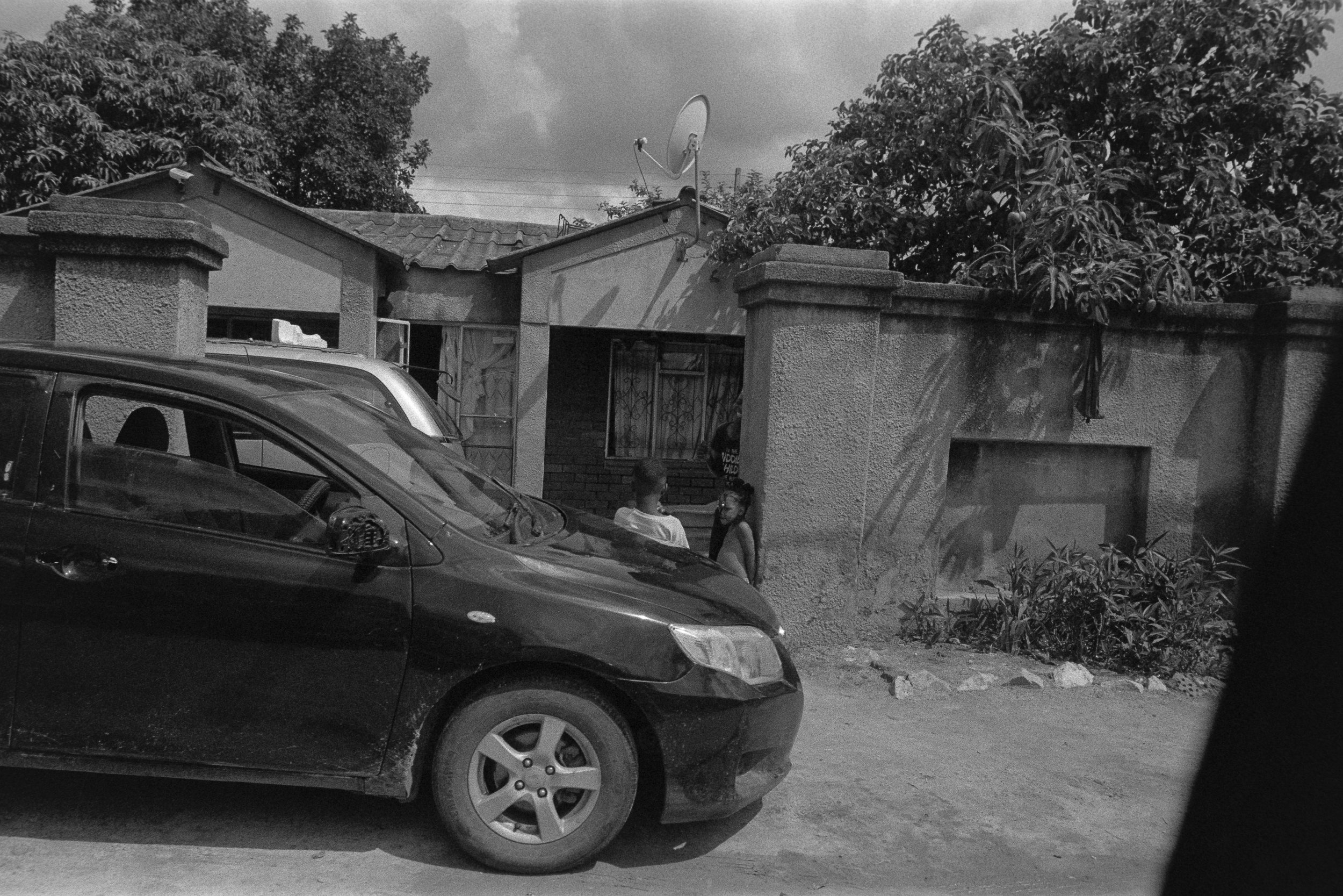 Black and white photo of two children standing and talking on a dirt street in front of a small house with a wall and gate. A black car is parked nearby, and a satellite dish is on the roof of the house. Trees and clouds are visible in the background