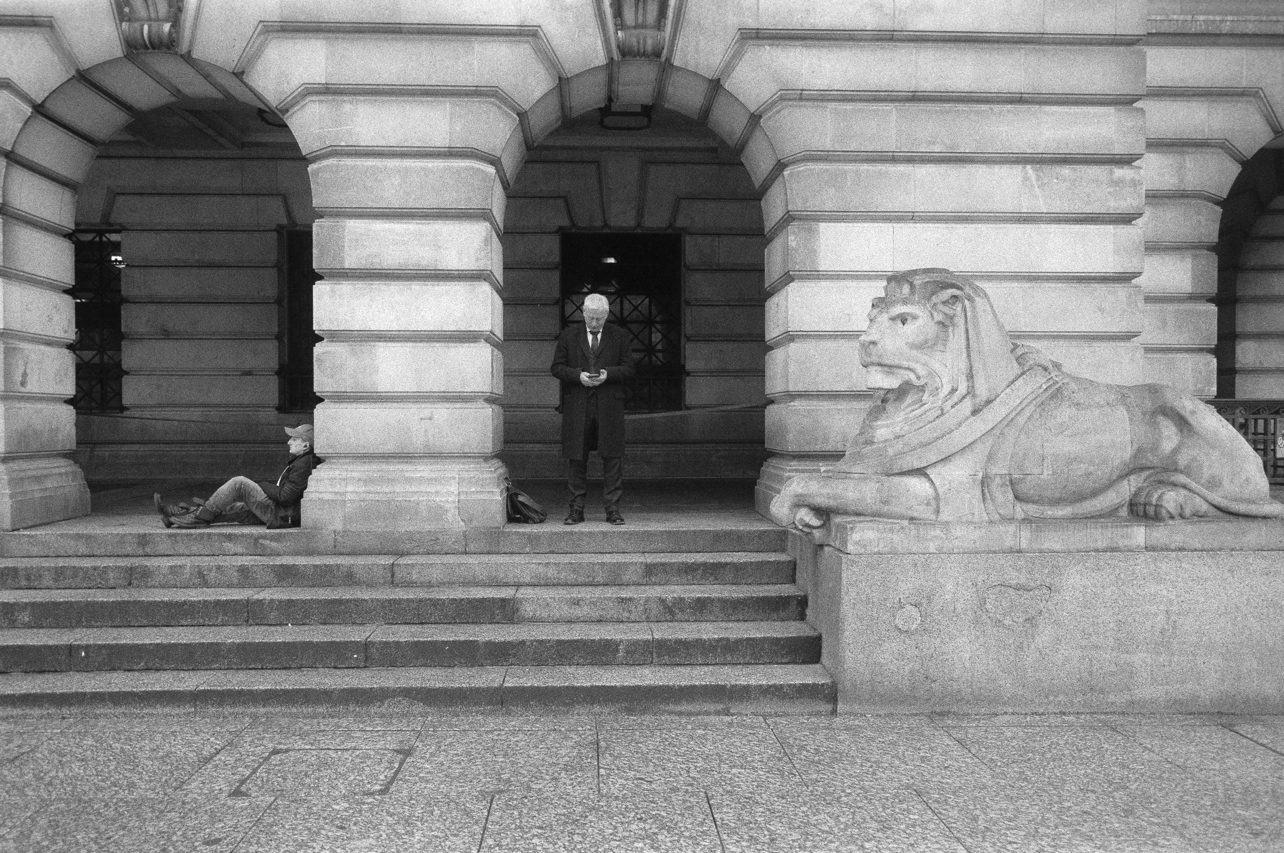 Black and white photo of an urban building with large stone steps, a lion sculpture on the right, a man sitting on the left, and a man standing in the center looking at his phone.