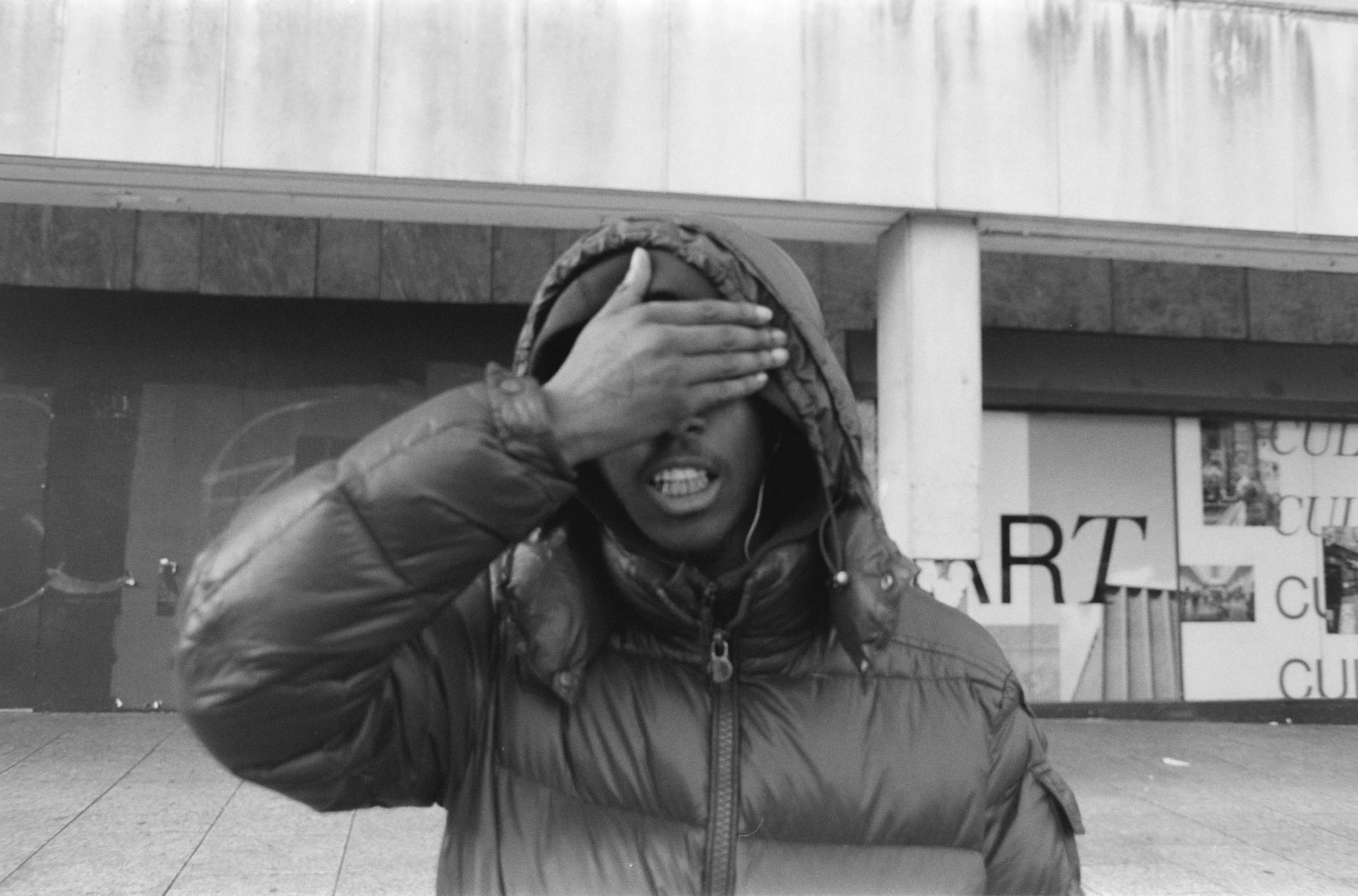 A person in a puffy jacket covering their eyes with one hand on an urban sidewalk.