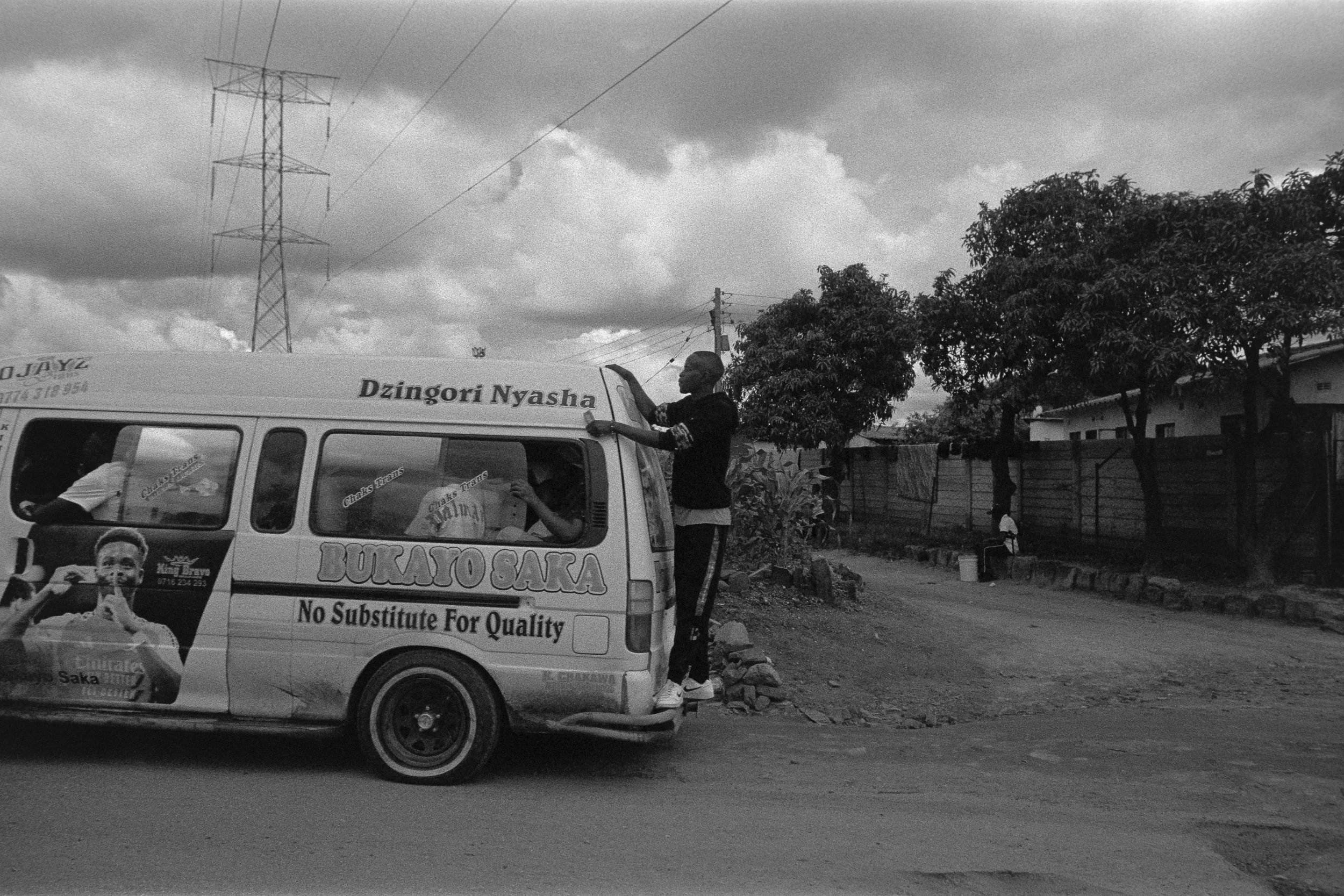 A small van decorated with Mr. Bukayo Saka's image and merchandise, parked on a dirt road. A person is standing on the back of the van, adjusting a sign while another person sits on the roadside. The scene includes trees, a fence, and an overcast sky