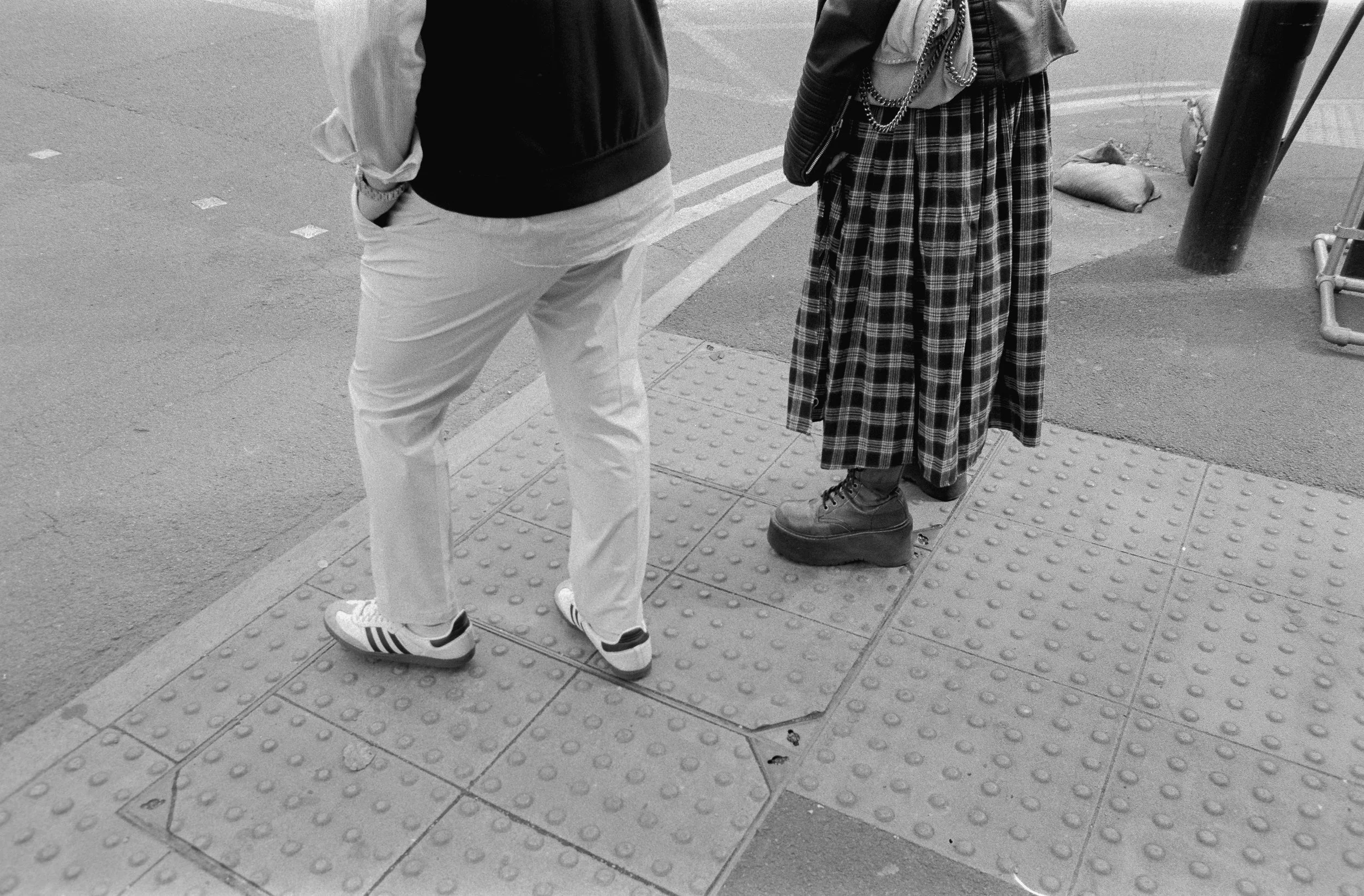Two people standing on a city sidewalk, one wearing white pants and sneakers, the other wearing plaid skirt, boots, and layered jewelry.