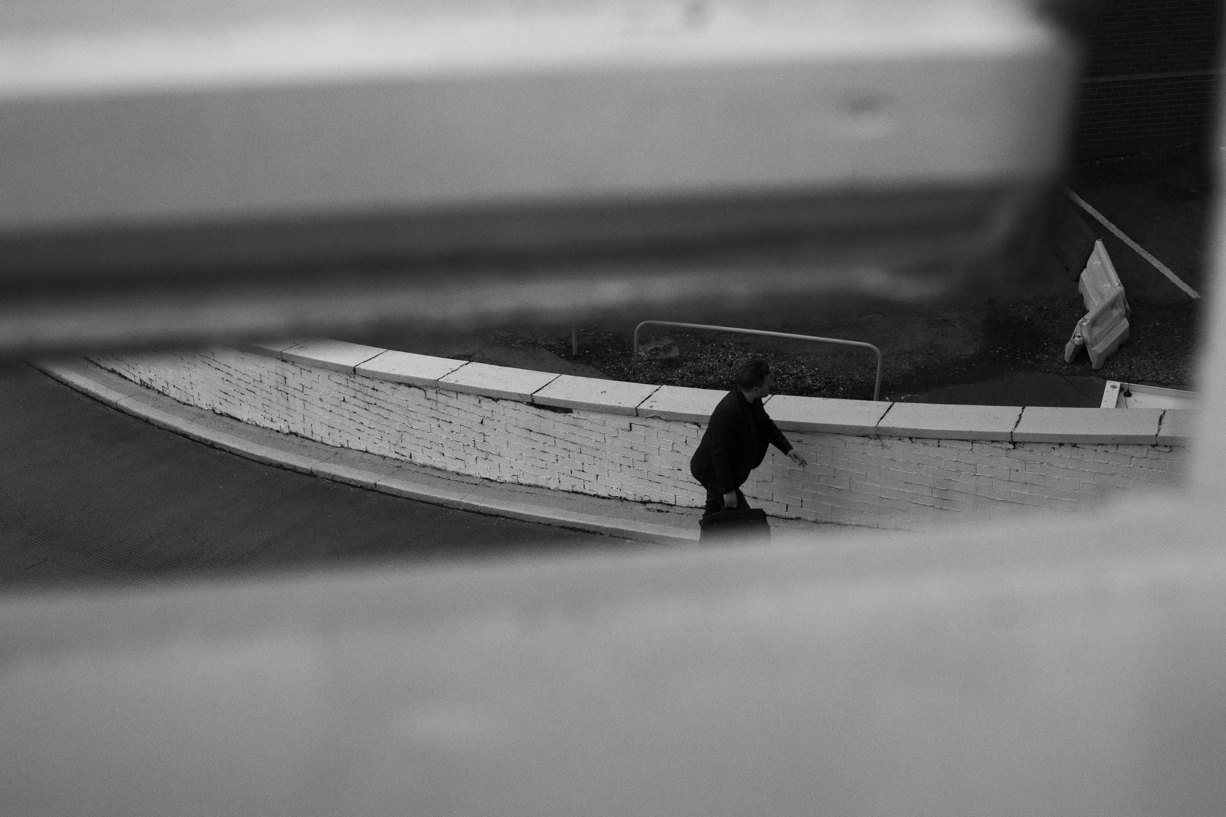 A person with glasses carrying a briefcase, walking along a white brick wall, viewed from above through horizontal blinds.