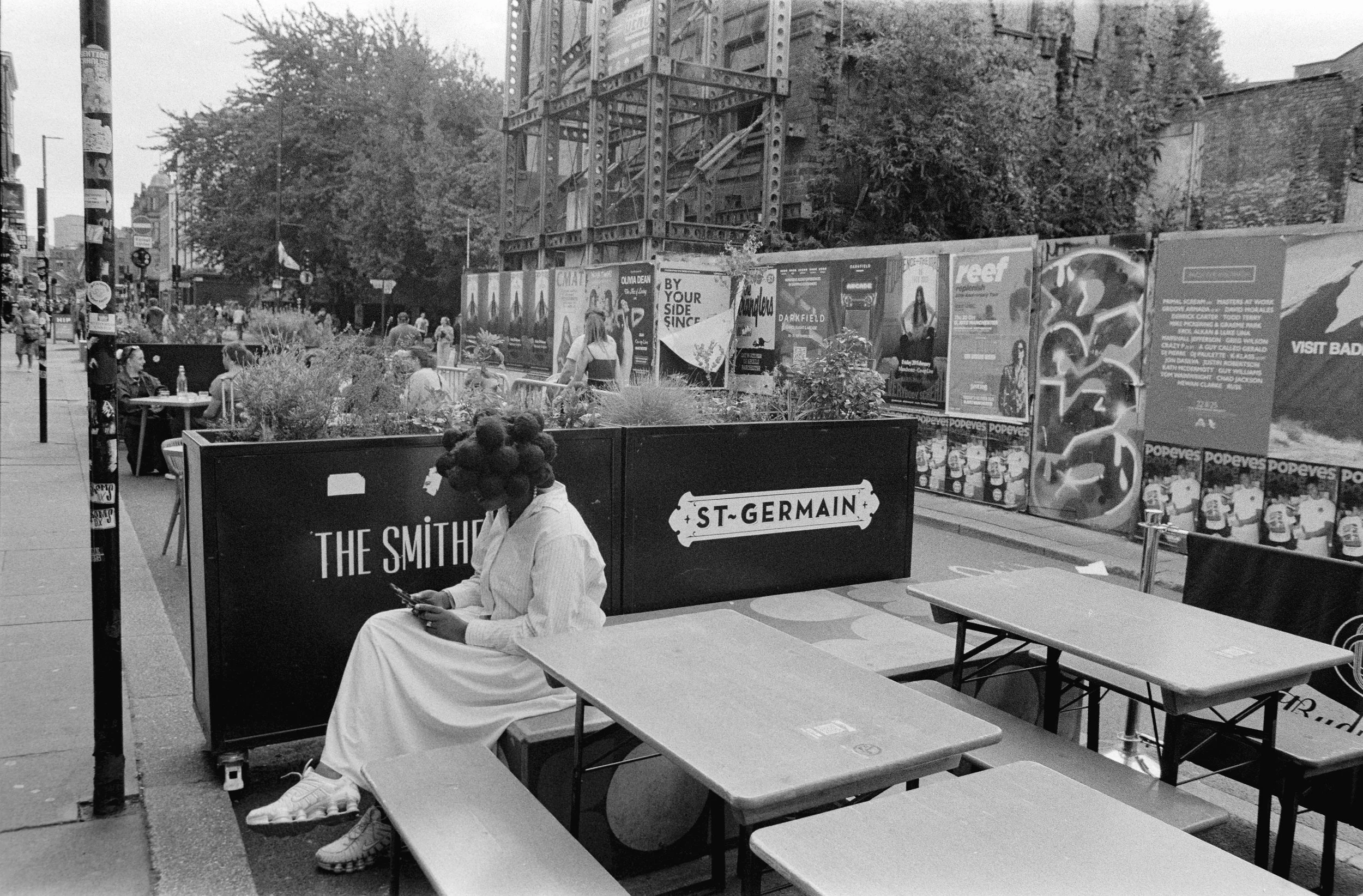A woman with hair styled in large curls sitting on a bench in an outdoor urban setting, looking at her phone. There are tables and chairs nearby, and a black wall with white text saying 'THE SMITH' and 'ST-GERMAIN'. In the background, there are sever