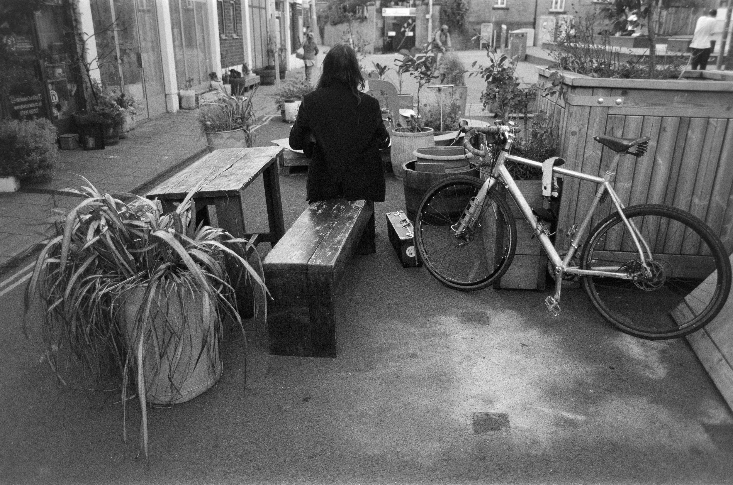 A woman sitting on a wooden bench at an outdoor cafe with potted plants and a bicycle parked nearby.