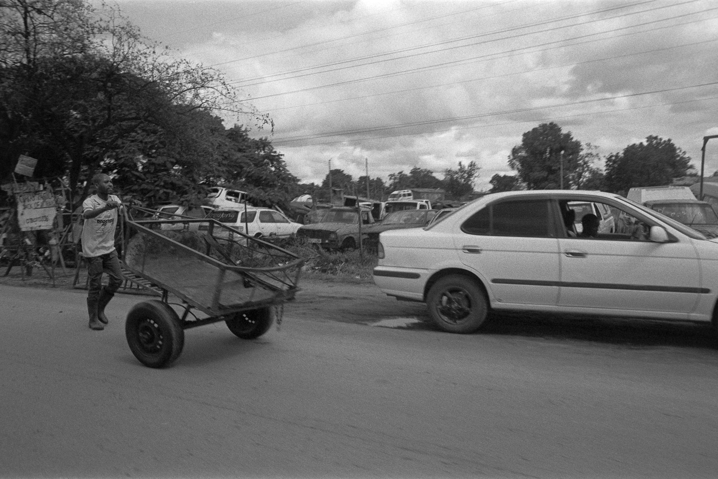 A man pulling a cart with a child sitting inside on a roadside, with cars parked and driving by, under cloudy skies.
